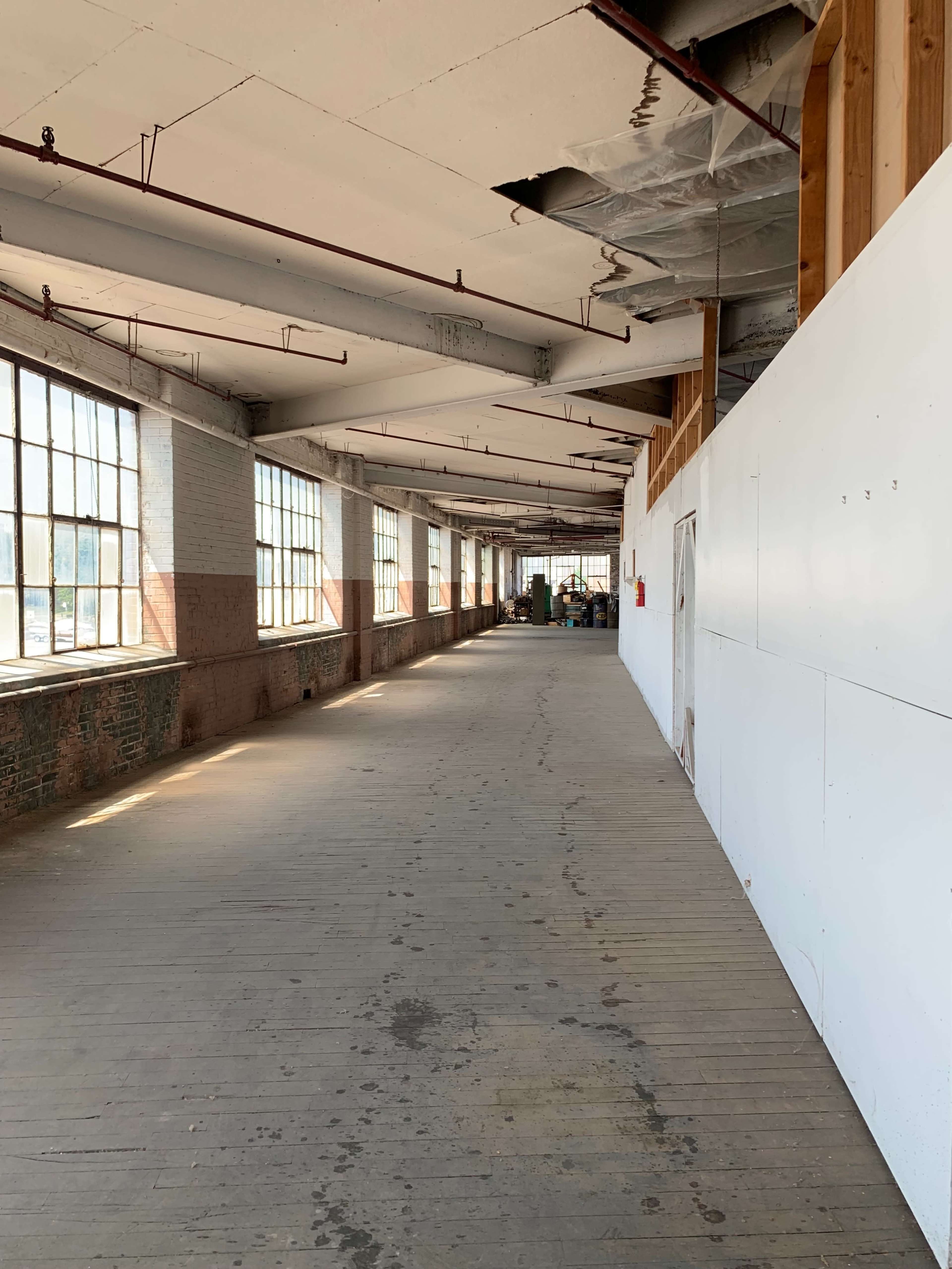 The image shows a long, empty corridor lined with large windows, featuring wooden floors and a white wall on one side.