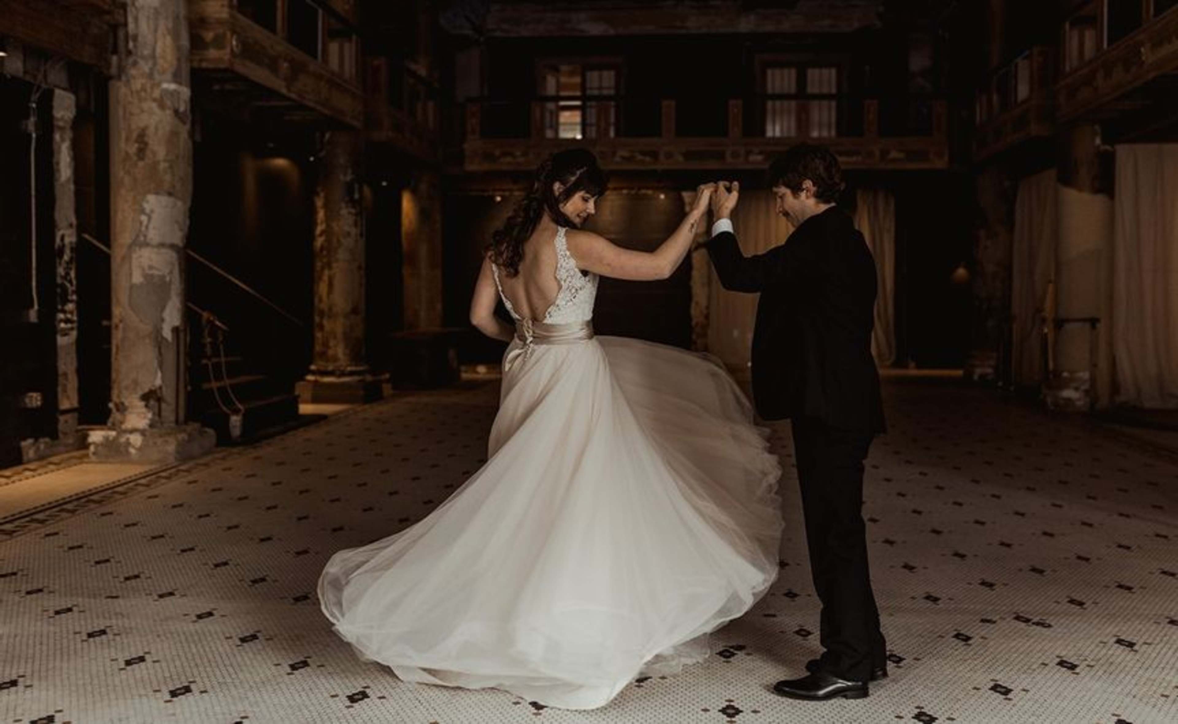 A couple dances in a spacious, dimly lit venue with a patterned tiled floor and exposed structural elements.