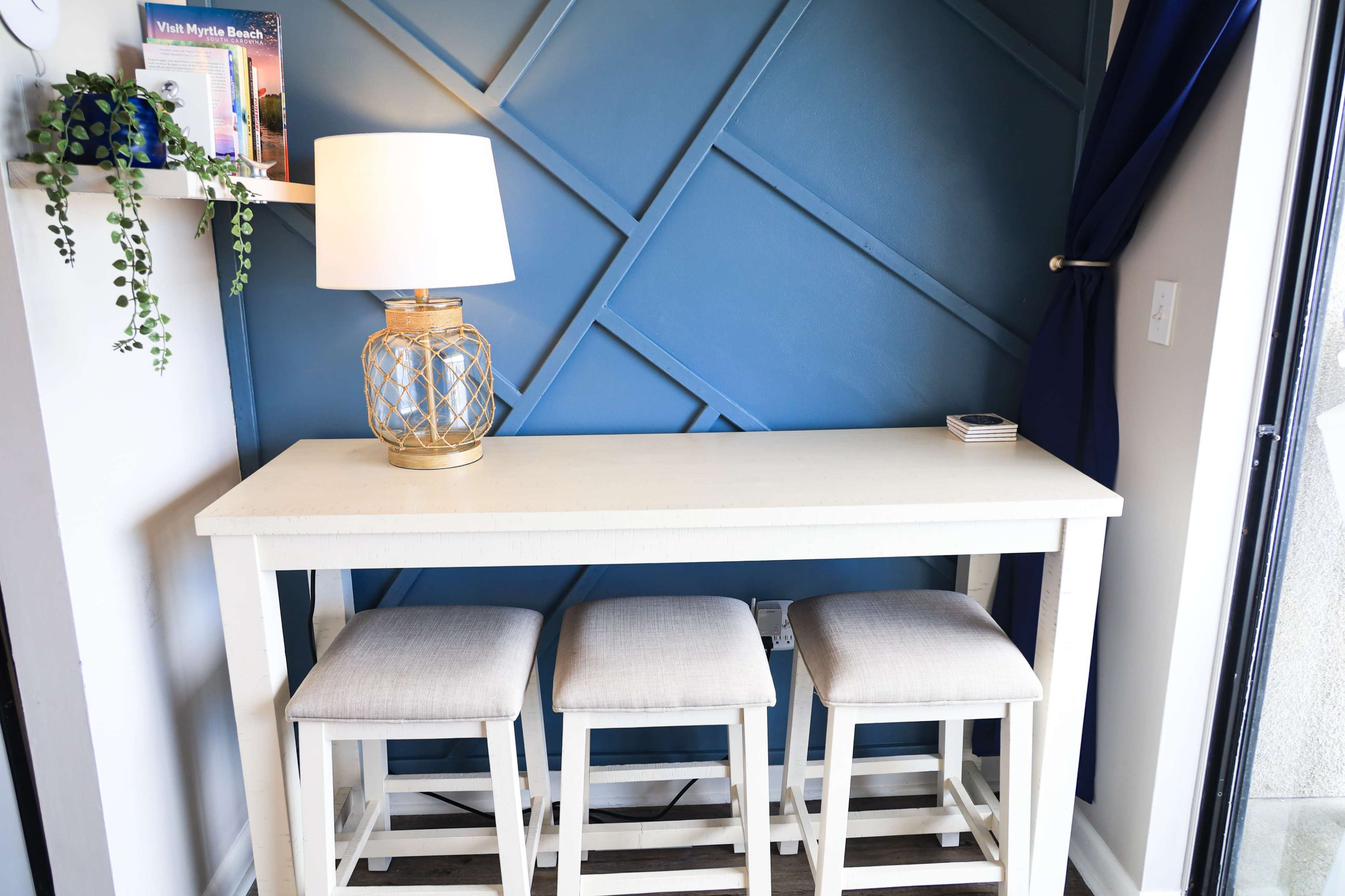 A small dining table with three stools is positioned against a blue accent wall, next to a lamp and a plant.