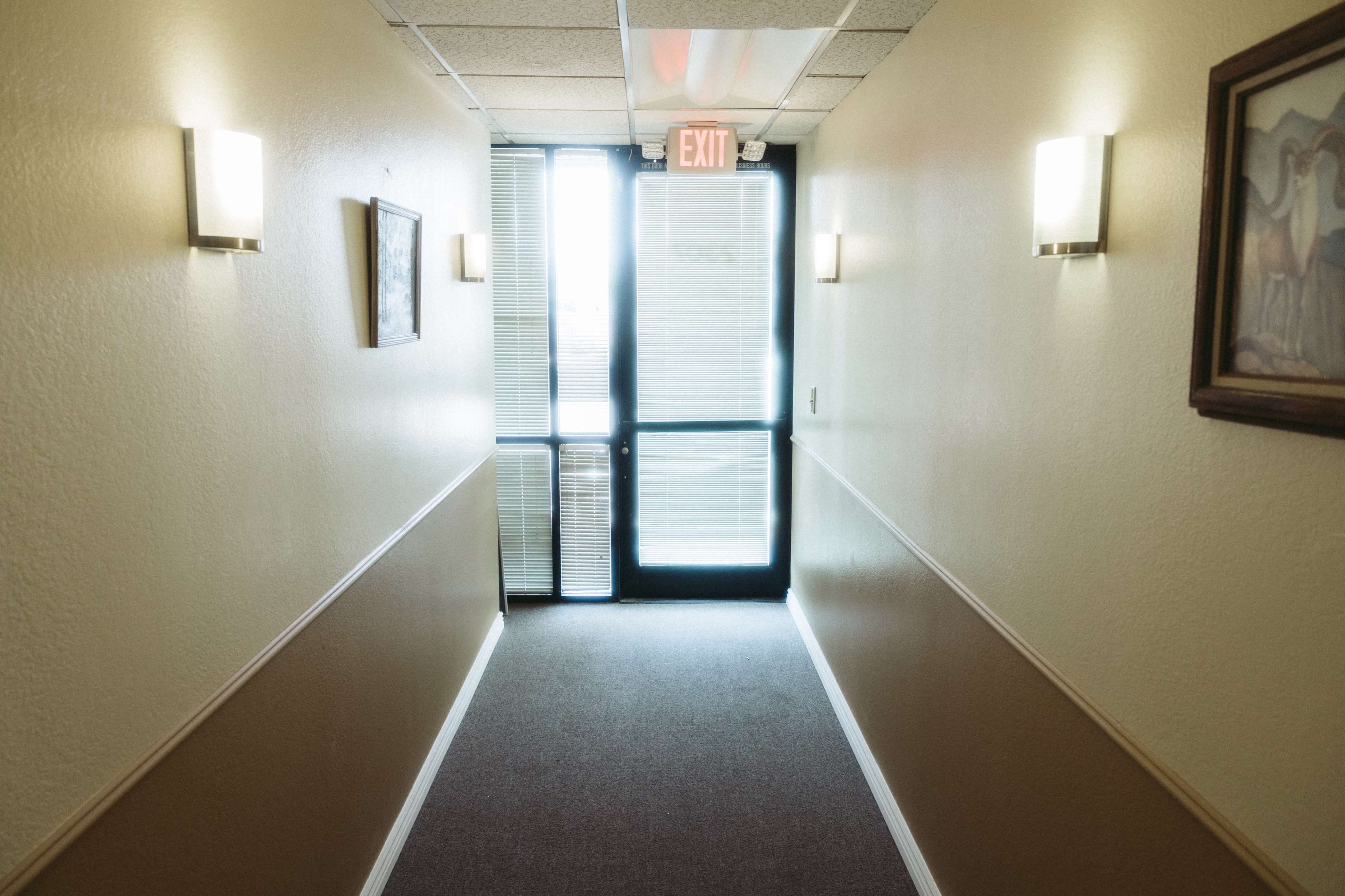 A brightly lit hallway leads to a glass exit door, with wall sconces illuminating the beige walls and a framed picture on the left.