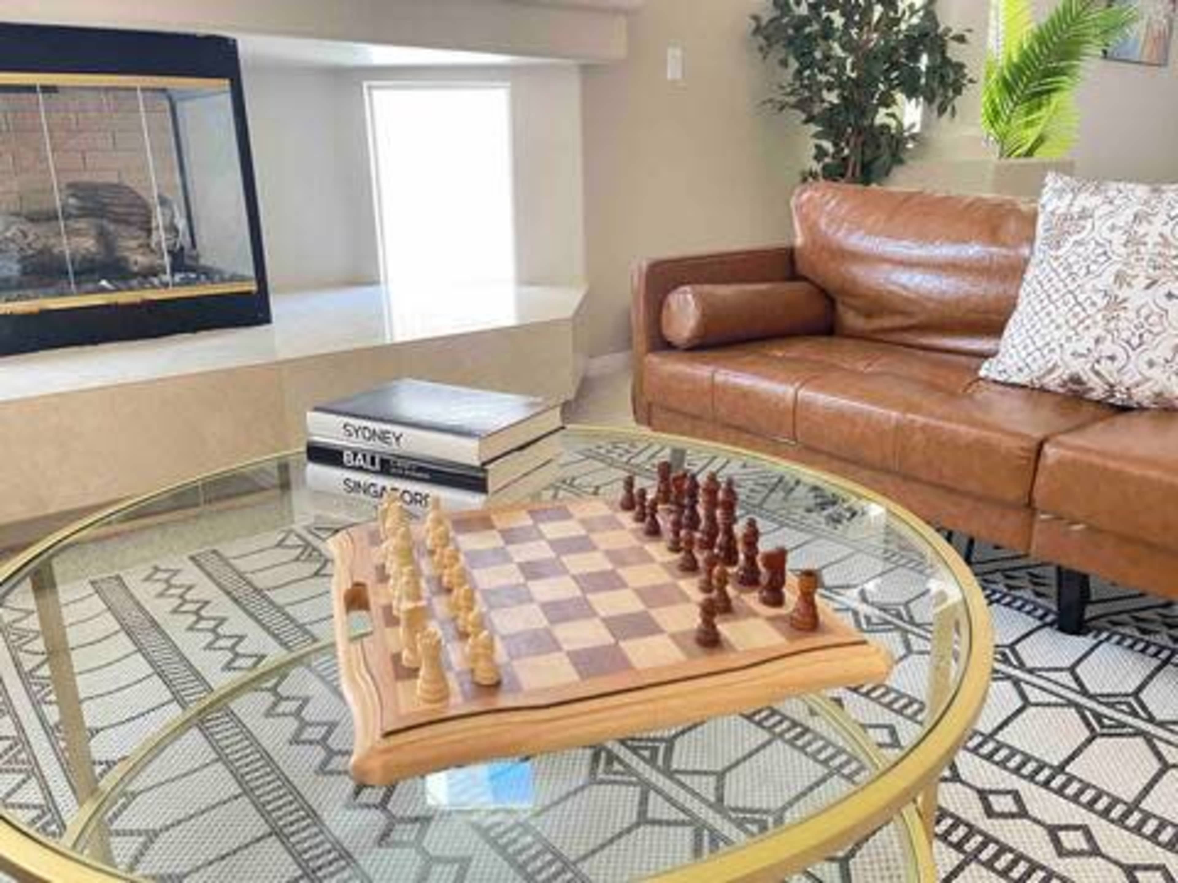 A chessboard with pieces set up for a game is placed on a glass coffee table in a living room featuring a leather couch and decorative plants.