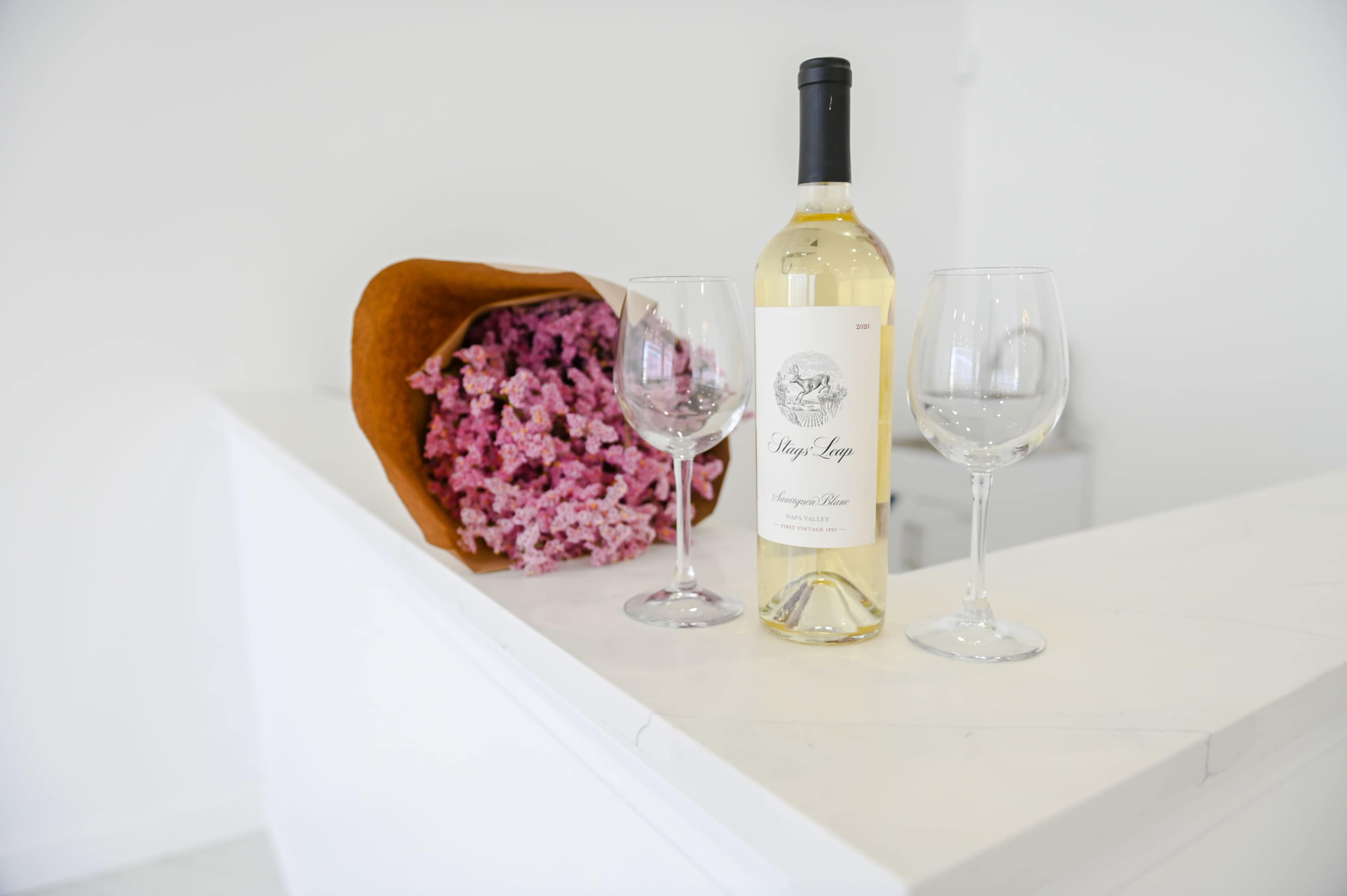 A bottle of white wine with two wine glasses and a bouquet of pink flowers are placed on a white countertop.