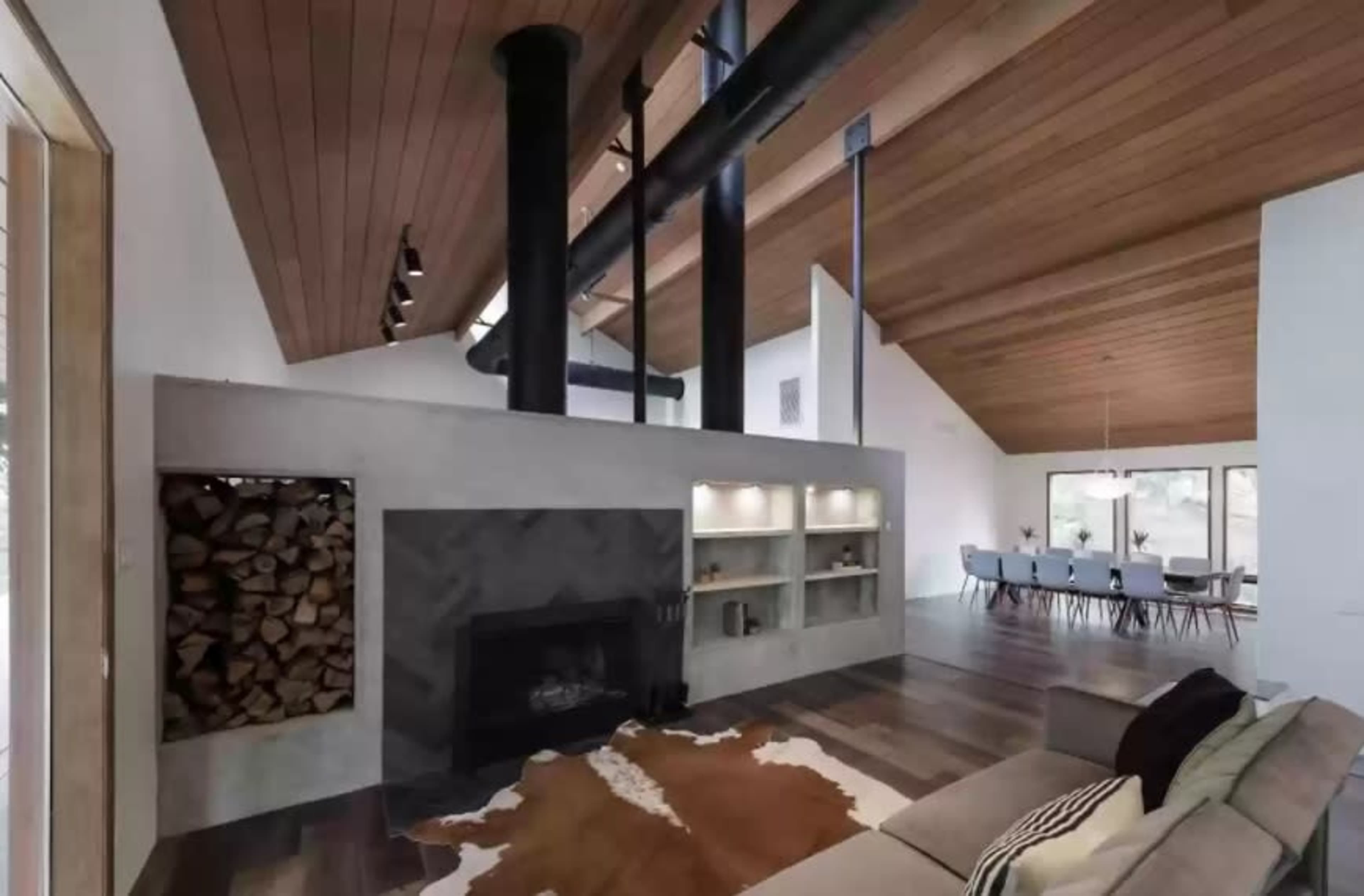 A modern living room featuring a fireplace with stacked wood, a sectional sofa, and a high vaulted ceiling with exposed beams.