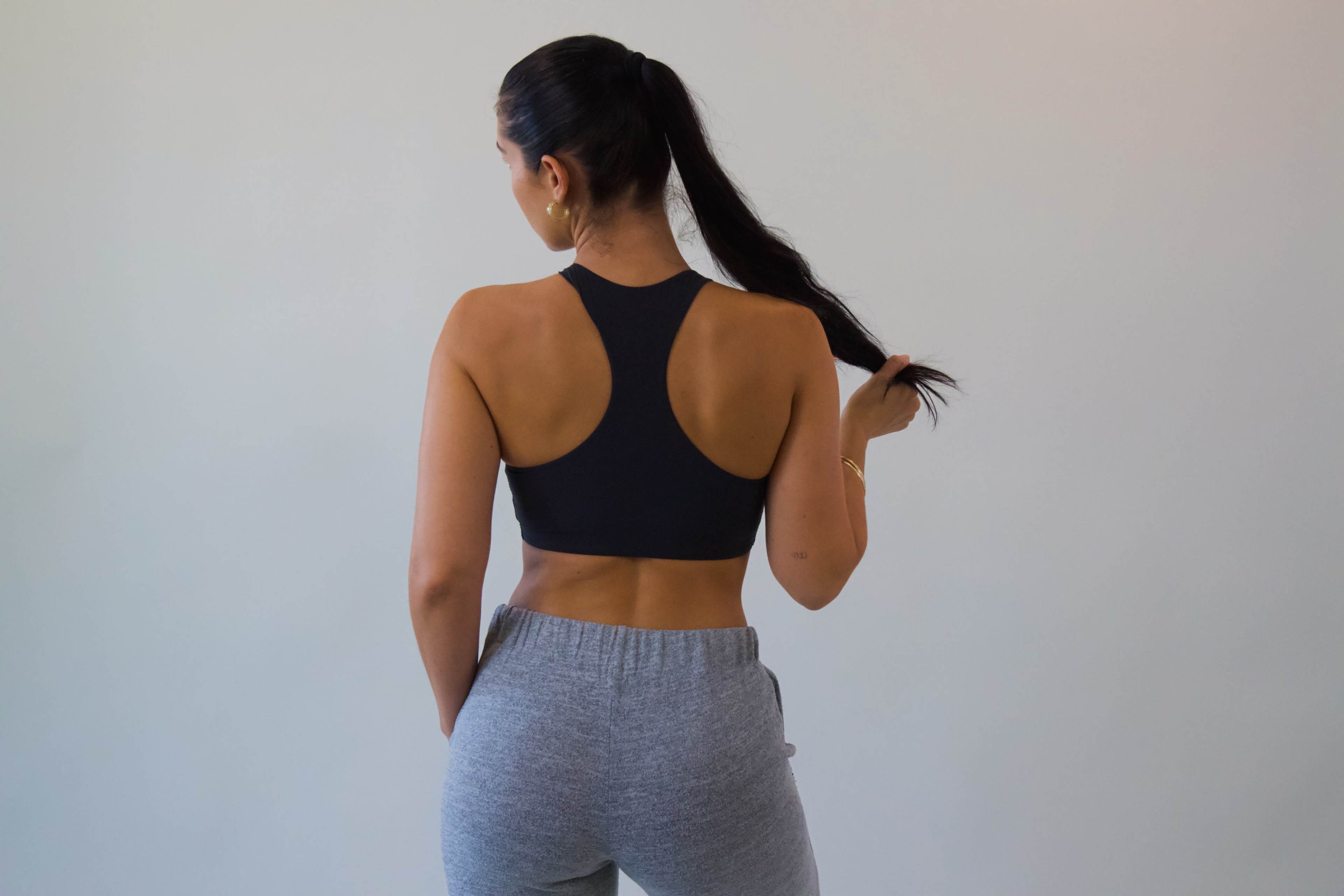 A woman in a black sports bra and gray sweatpants stands with her back to the camera, holding her long hair.