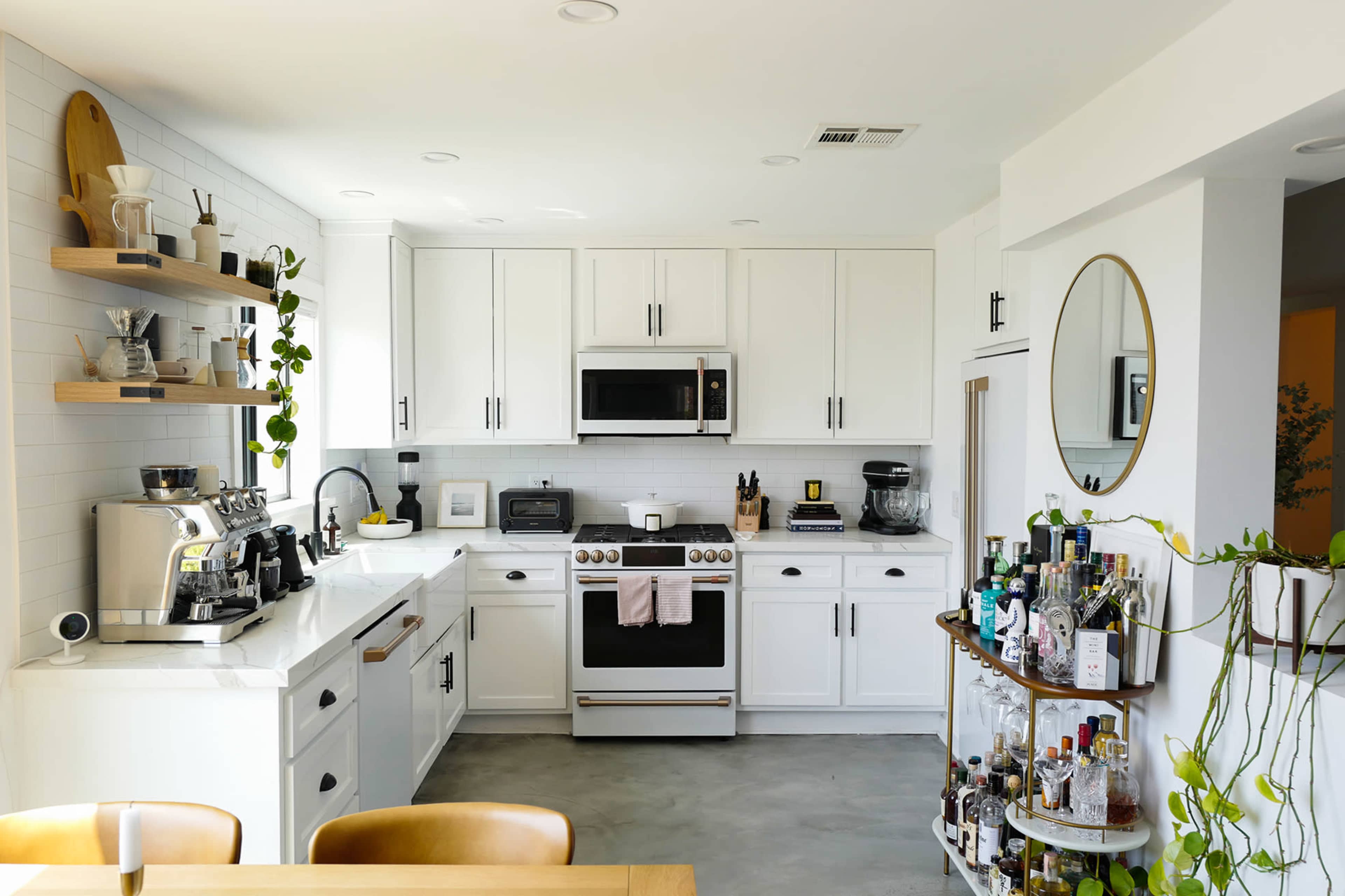 A modern kitchen features white cabinetry, stainless steel appliances, a countertop with coffee equipment, and a circular mirror above a bar cart filled with various bottles.