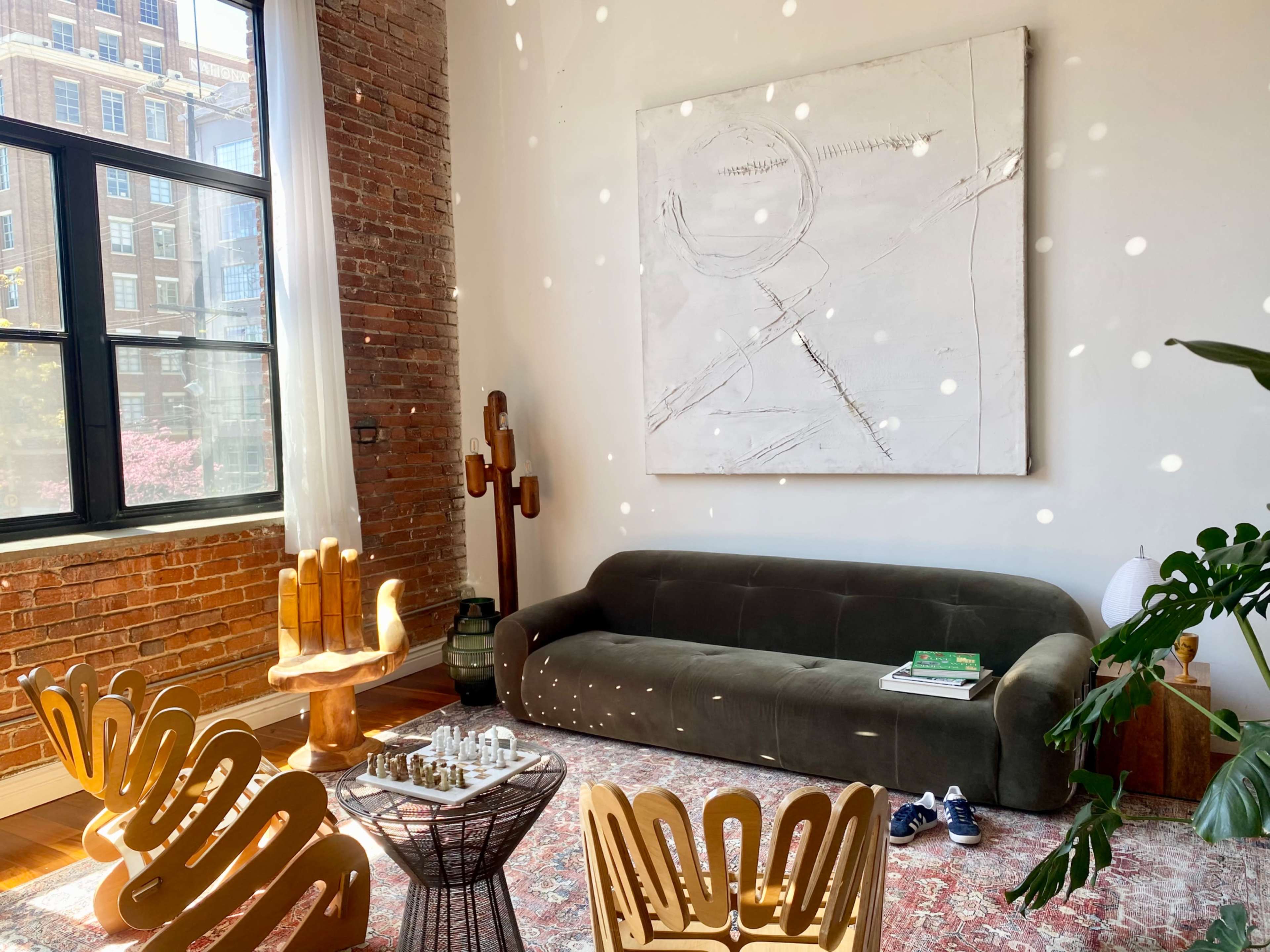 A modern living room with a green sofa, a large abstract painting on the wall, and several wooden chairs arranged around a coffee table with a chess set.