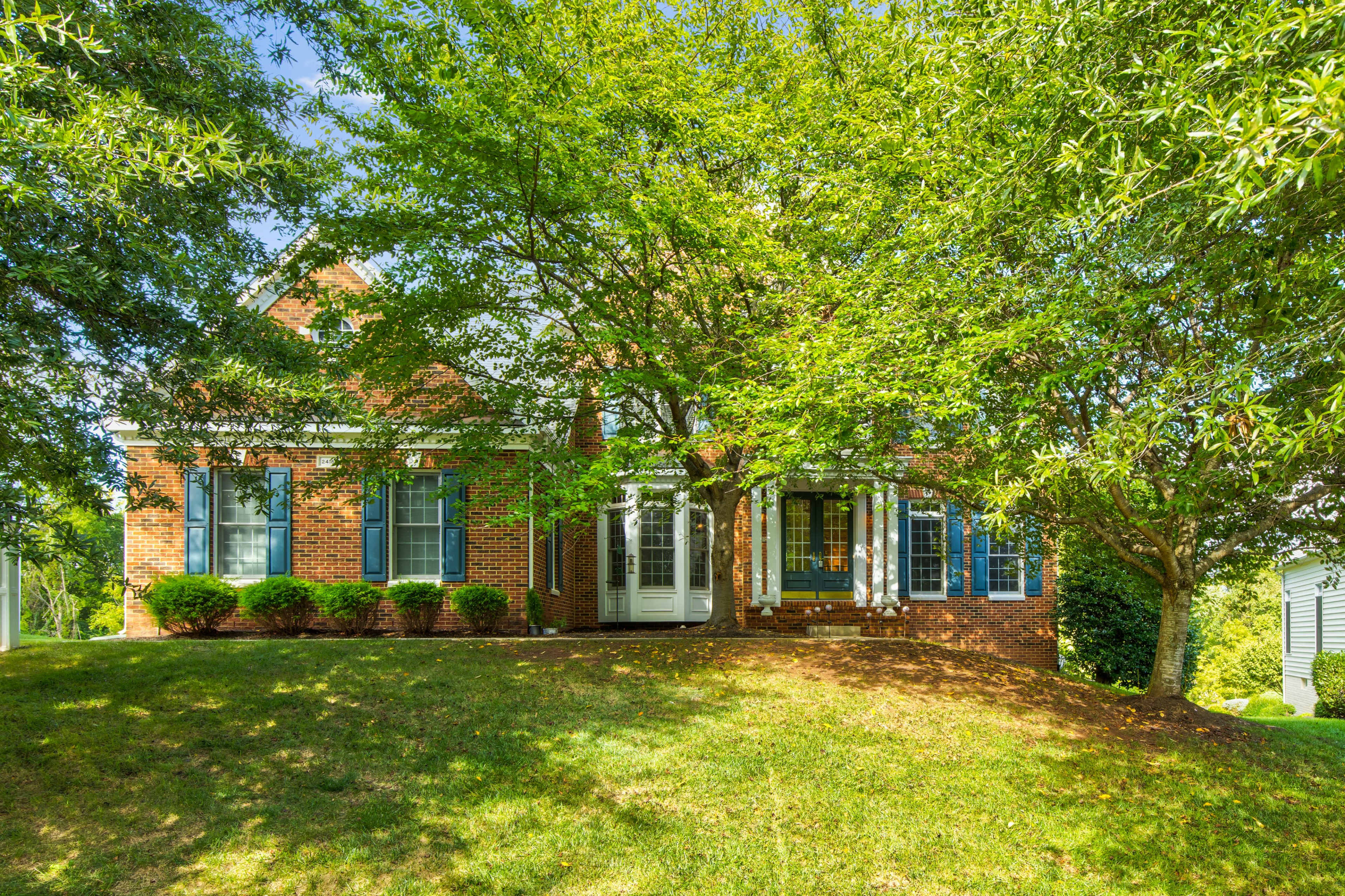 A brick house with blue window shutters is situated on a sloped lawn surrounded by trees.
