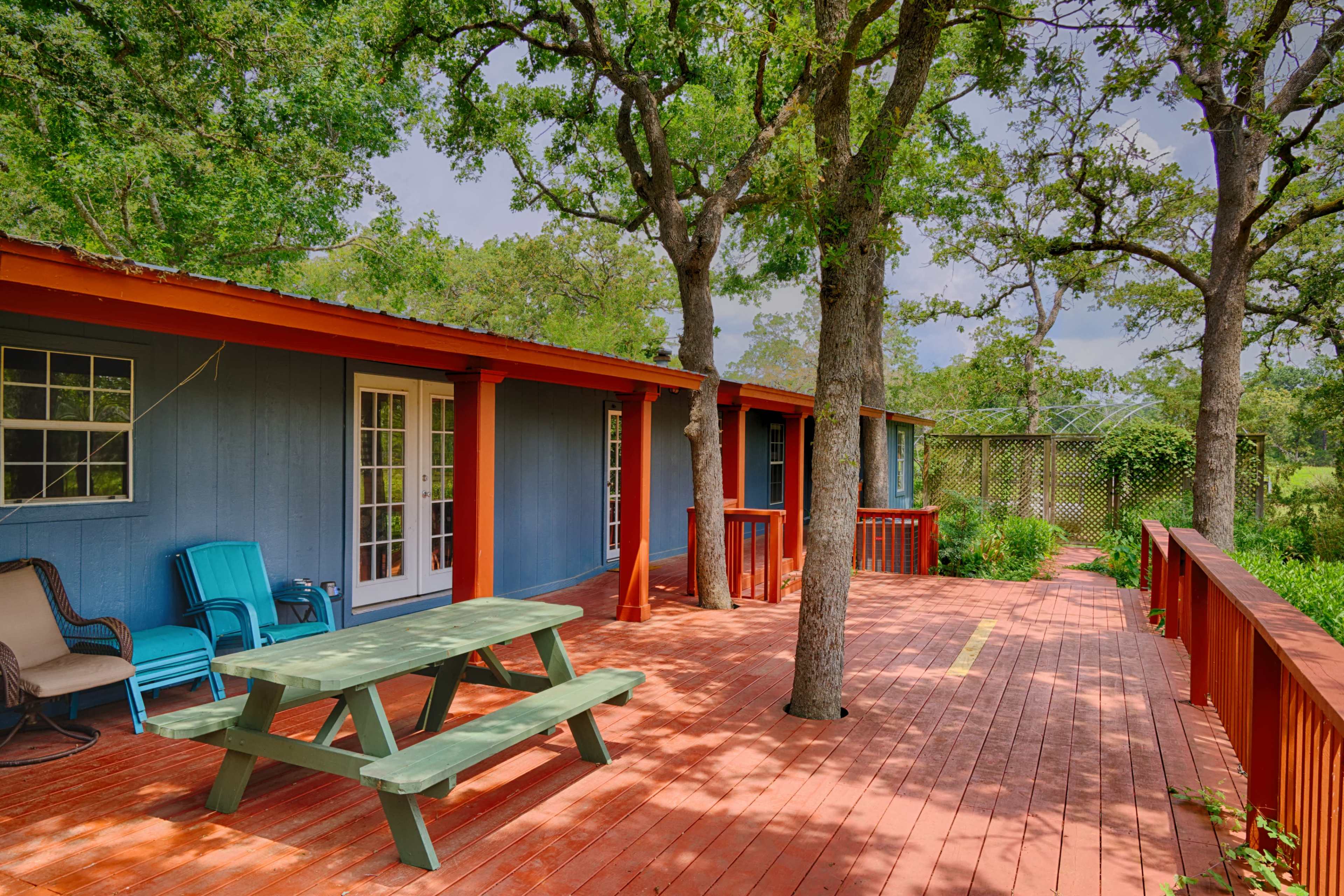 A wooden deck with a picnic table and chairs in front of a blue building surrounded by trees.