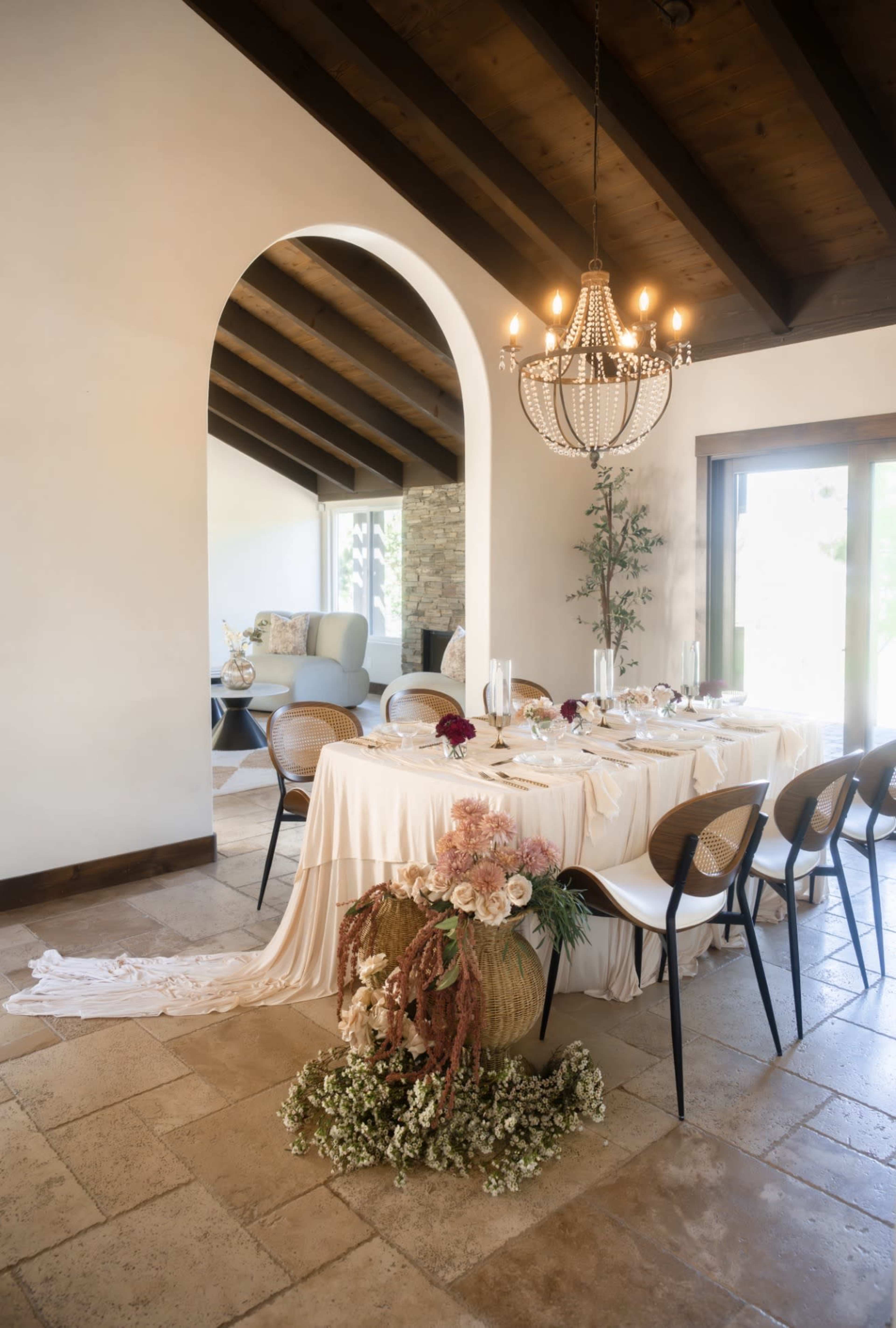 A dining table adorned with a tablecloth and floral arrangements is set beneath a chandelier in a room featuring wooden beams and an arched entryway.