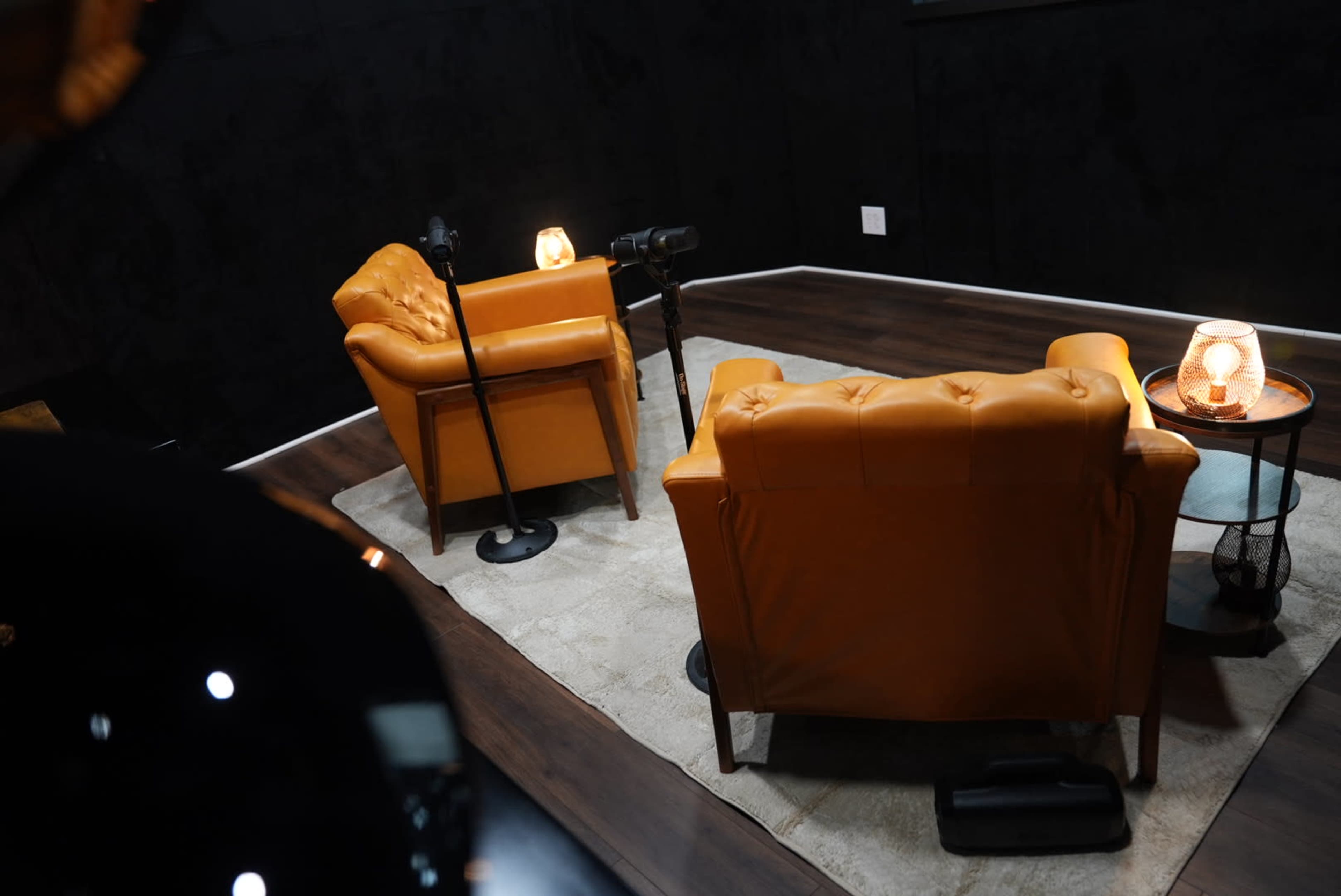 The image shows two orange armchairs positioned across from each other on a gray rug, with a small table and a lamp between them in a dimly lit room.