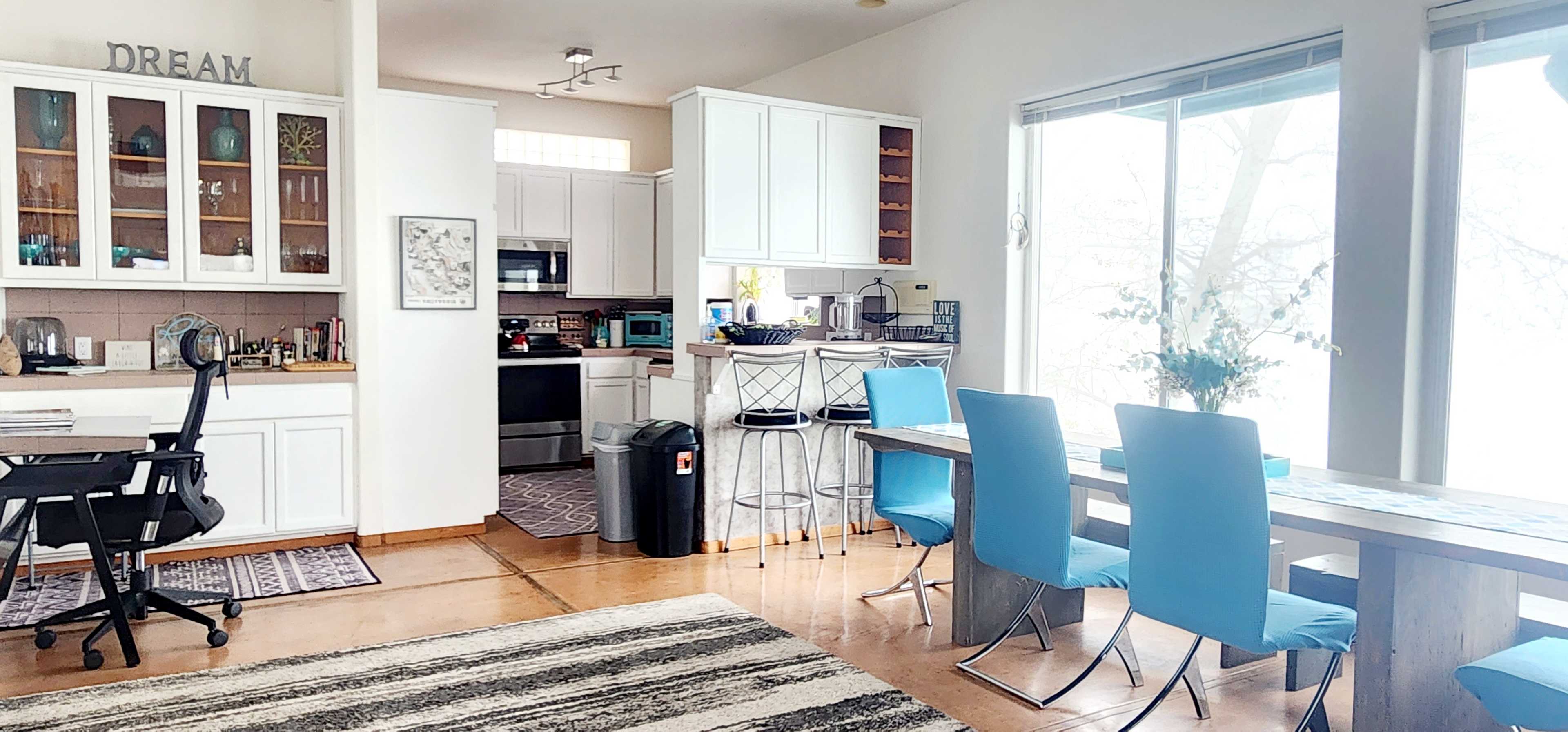 The image shows a modern, open-concept kitchen and dining area with a wooden table, blue chairs, and large windows allowing natural light to enter.