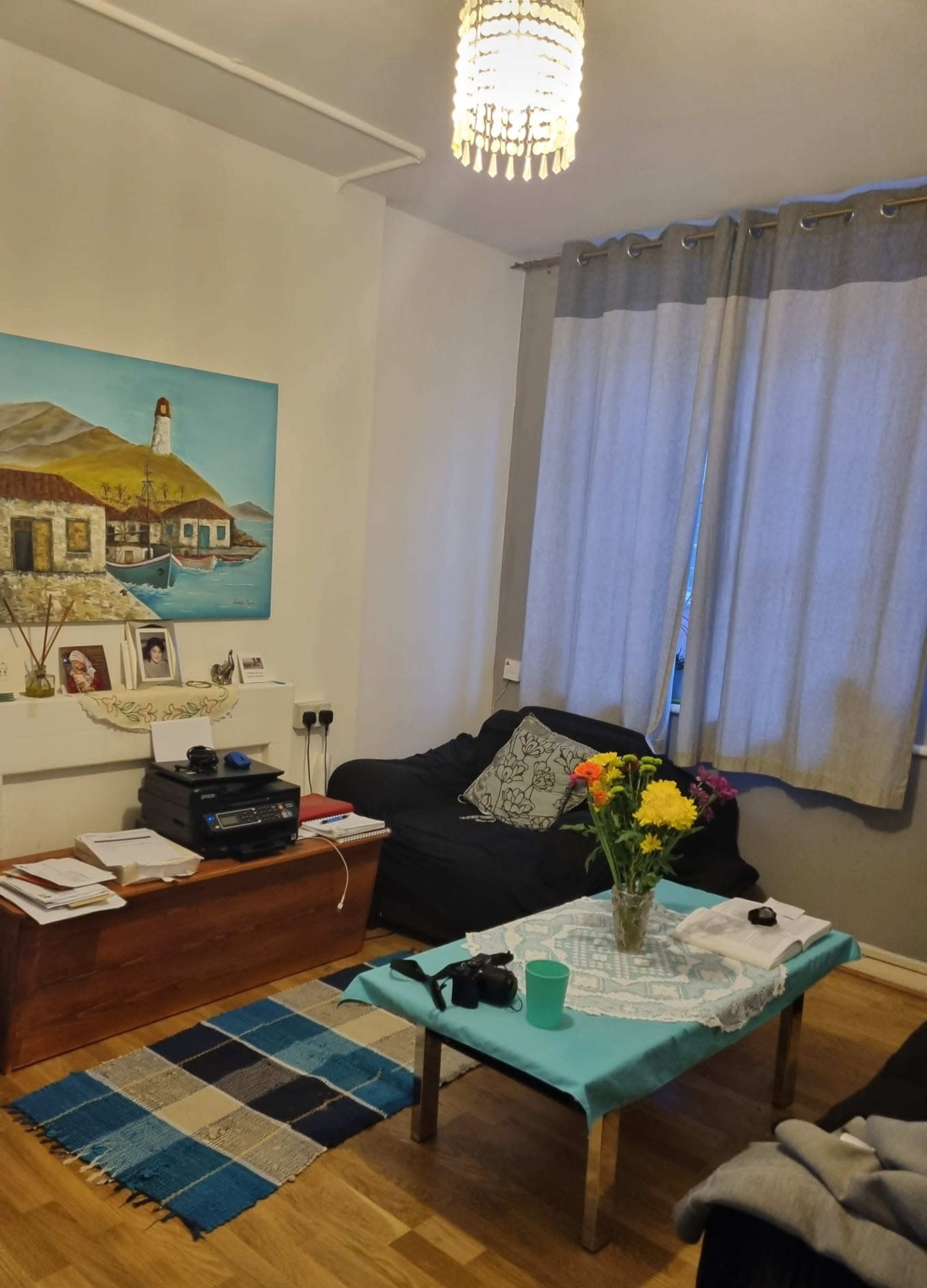A cozy living room with a blue and white theme, featuring a wooden coffee table, a dark couch, a colorful painting on the wall, and a bouquet of flowers in a vase.