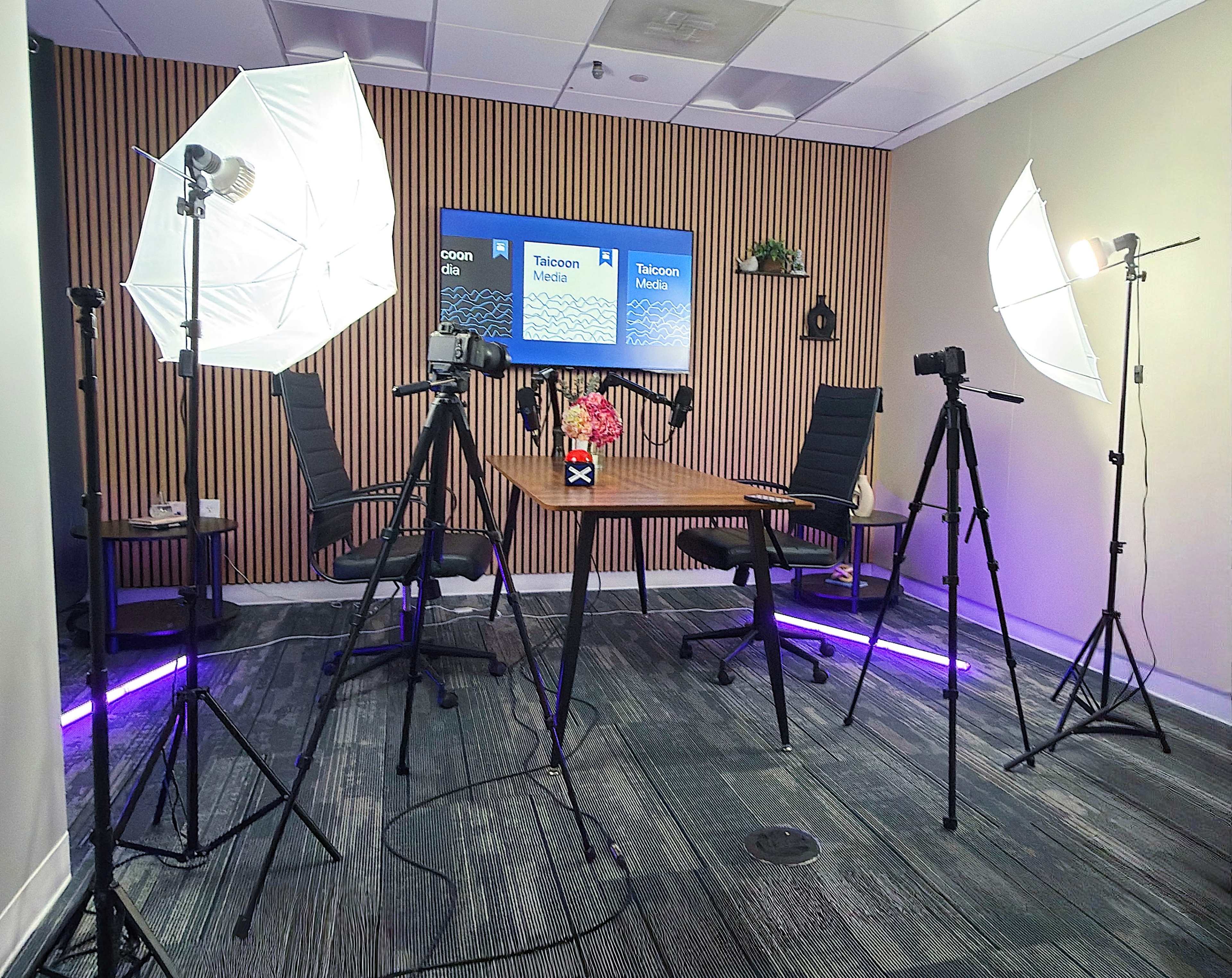 The image shows a well-organized studio setup with two cameras, softbox lights, a wooden table adorned with flowers, and two empty chairs in front of a backdrop displaying the "Takeson Media" logo.