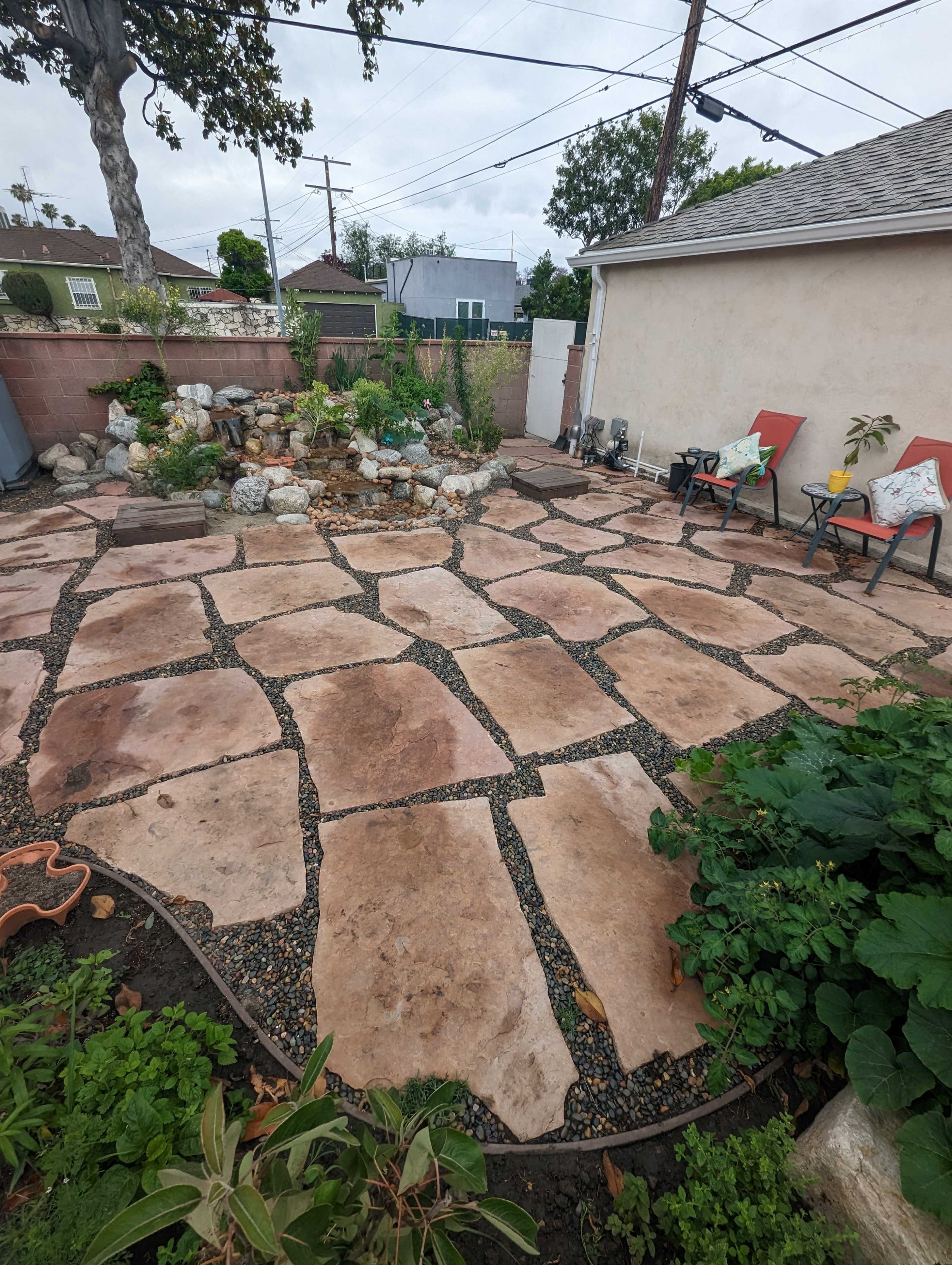 The image shows a stone patio with large flat stones arranged over gravel, surrounded by greenery and a rock feature suggesting a water element in a backyard setting.