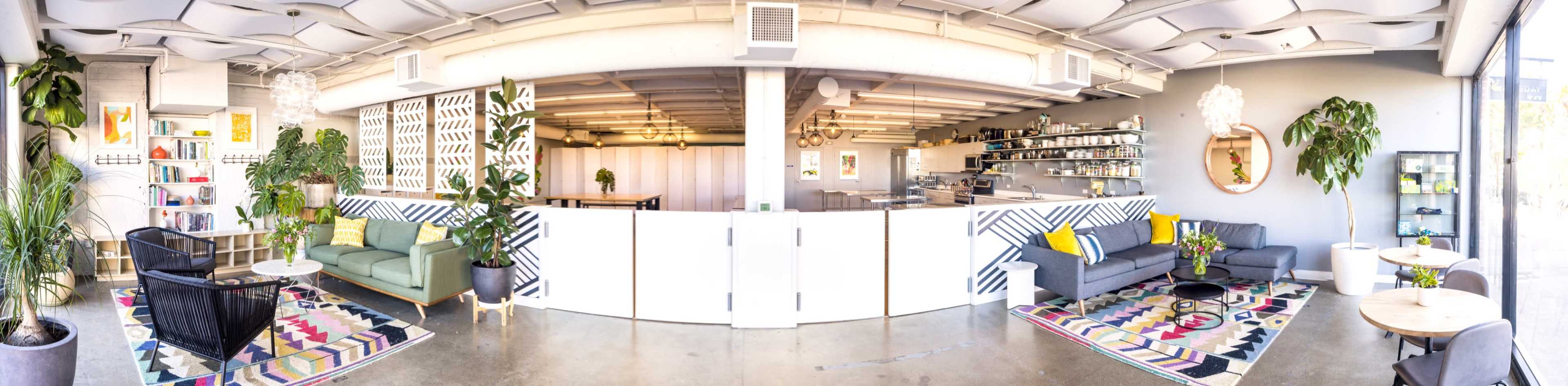 A spacious, brightly lit lounge area with various seating arrangements, plants, and a modern decor featuring patterned rugs and shelves filled with items.