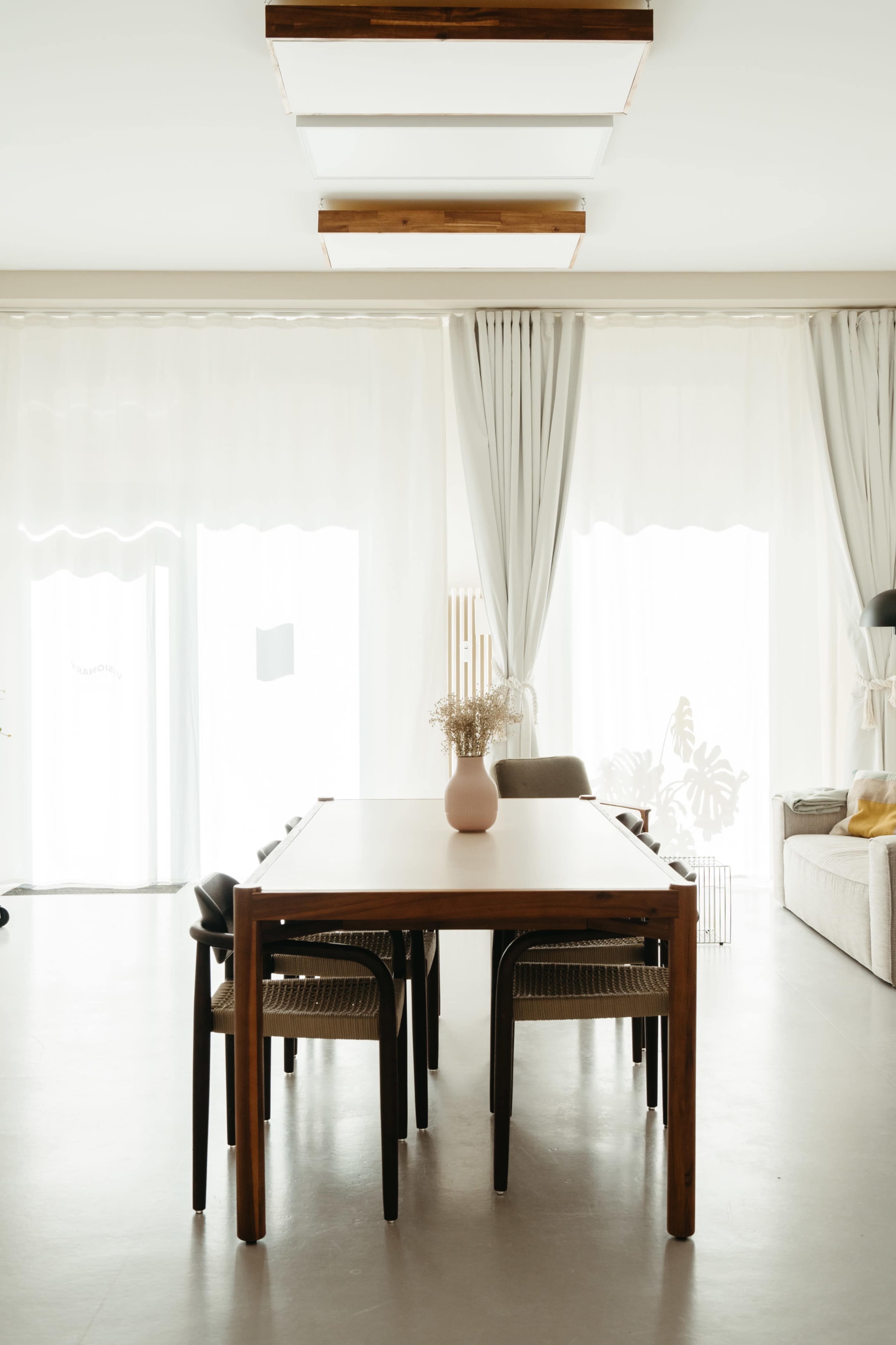 A modern dining room features a large wooden table surrounded by several chairs, with white curtains and natural light streaming in through the windows.
