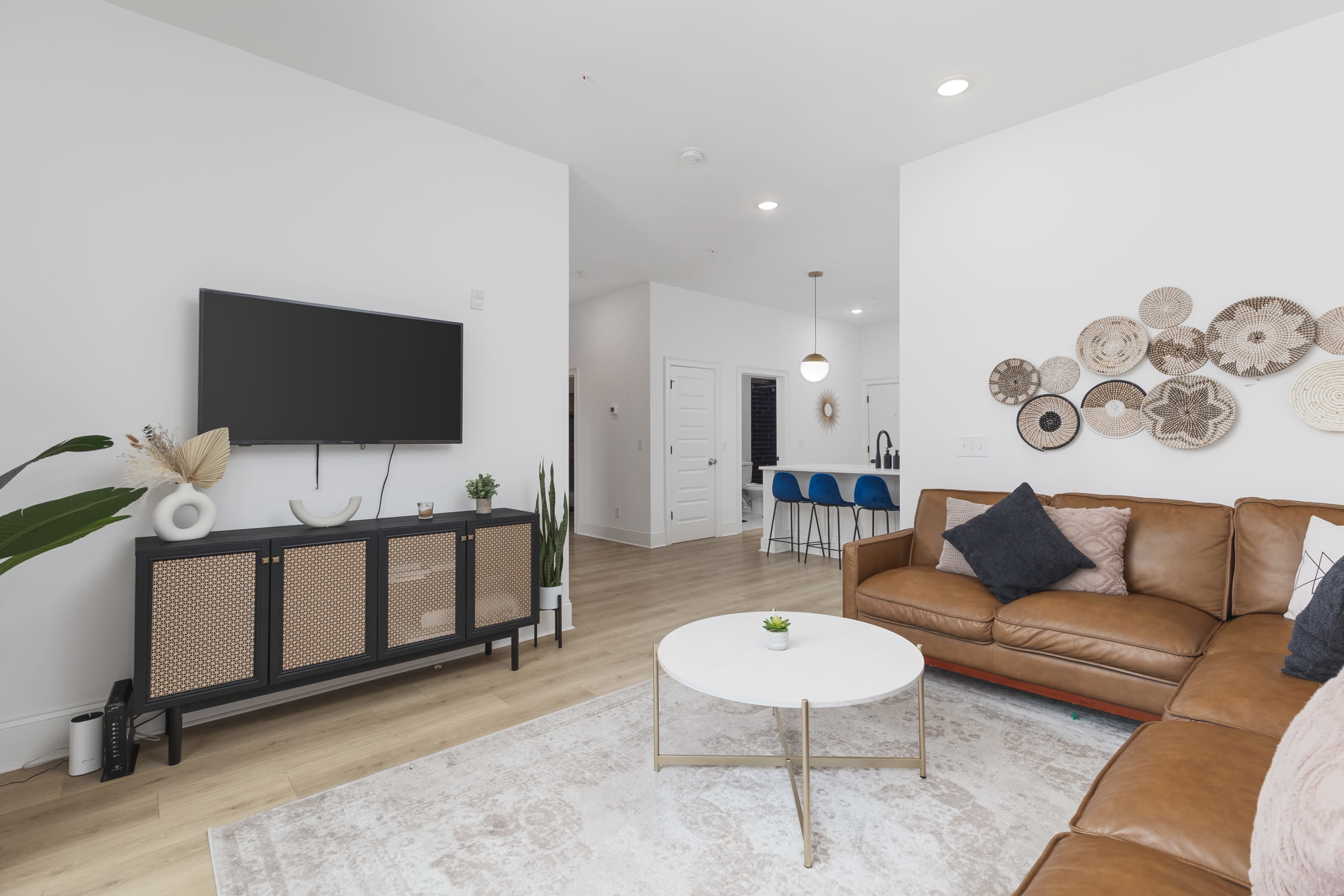 A modern living room features a brown leather sofa, a round coffee table, a television on a console unit, and decorative woven wall art.