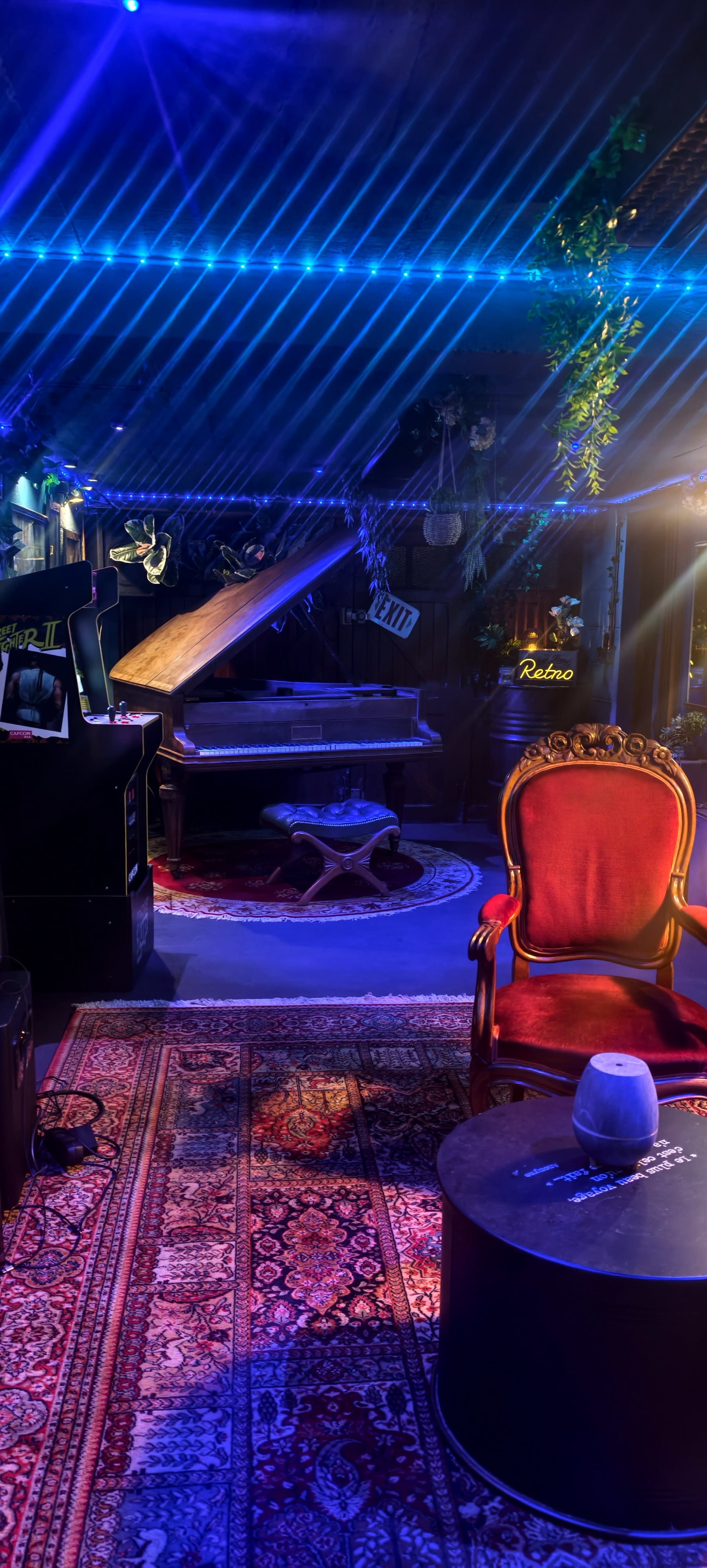 The image depicts a cozy room with a vintage piano, an arcade machine, a plush red chair, and a small table, all illuminated by colorful lights and surrounded by greenery.