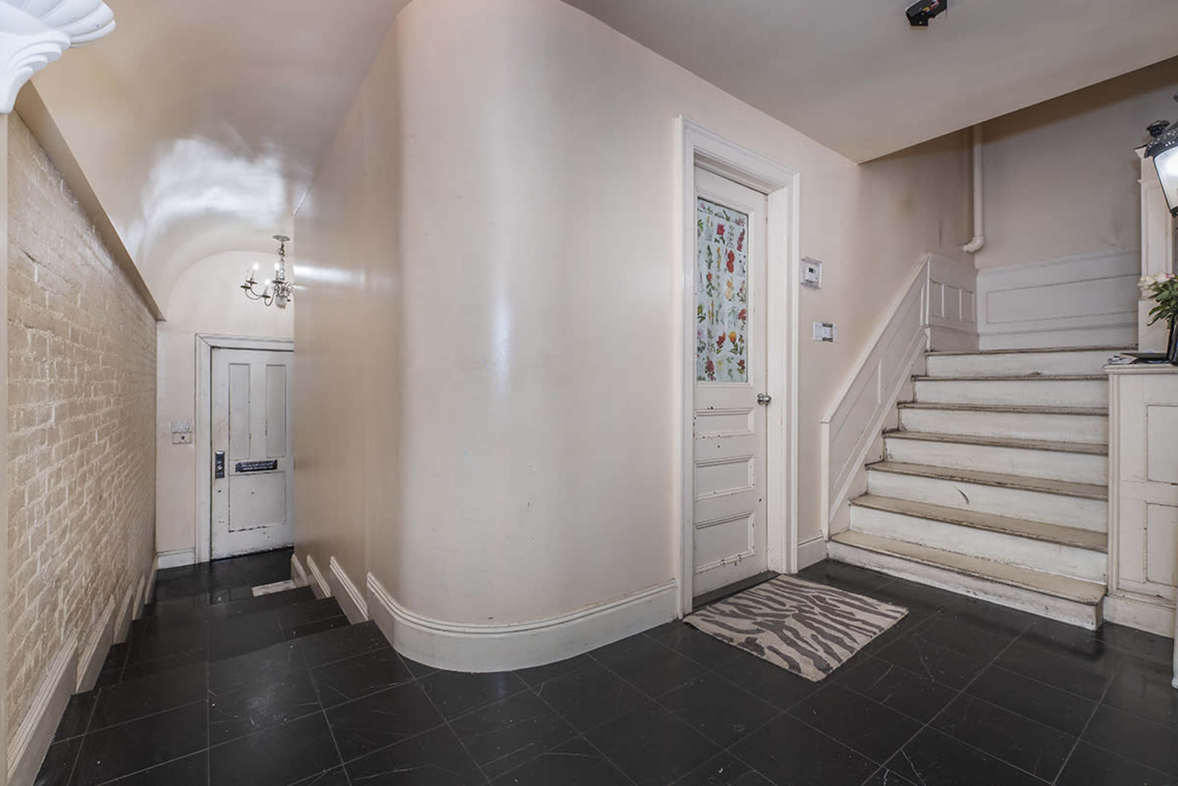 The image shows a hallway with a staircase on the right, a door near the bottom of the stairs, and a patterned runner on the black tiled floor.