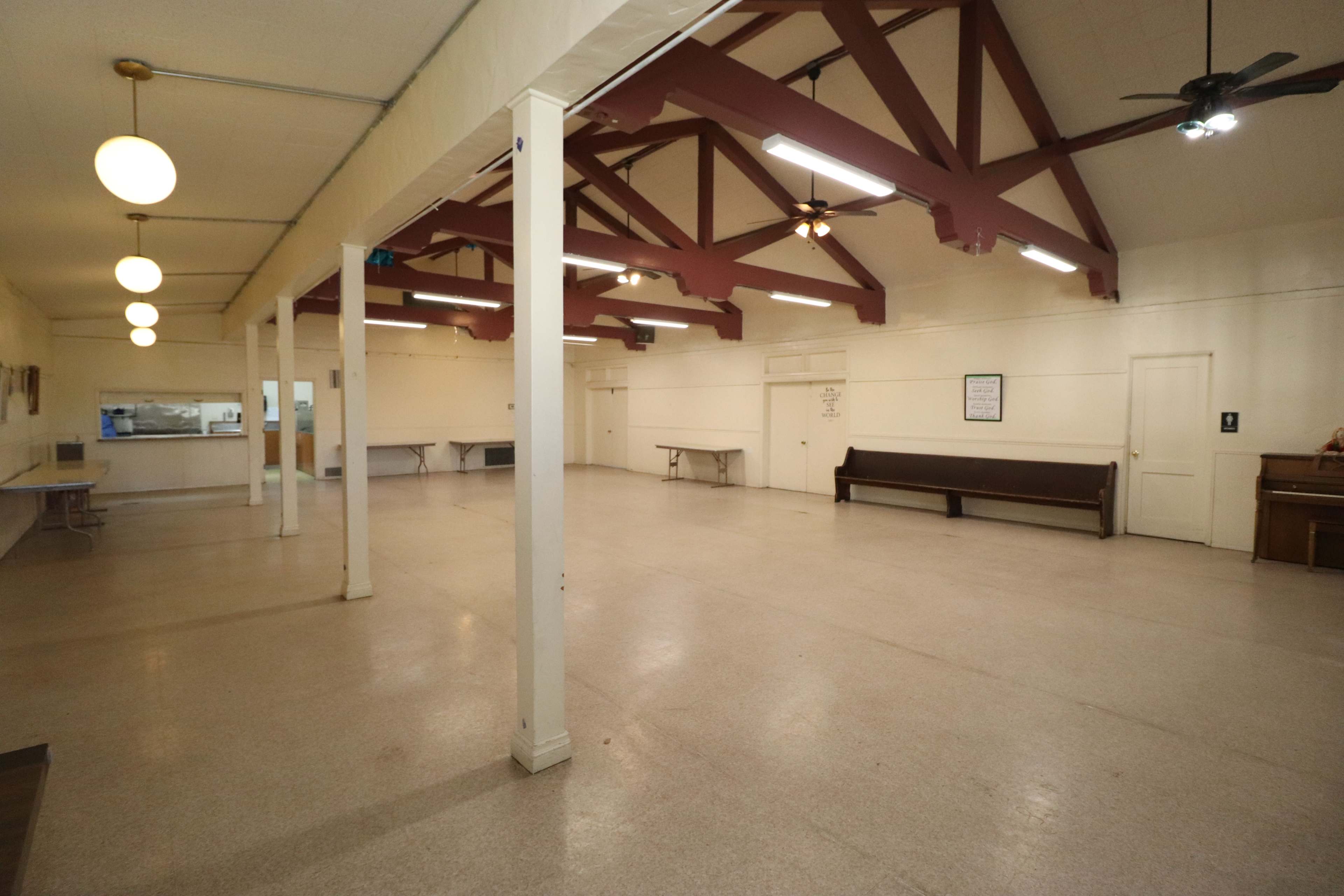 A spacious hall with high ceilings, wooden beams, and several tables arranged along the sides, as well as a serving area visible in the background.