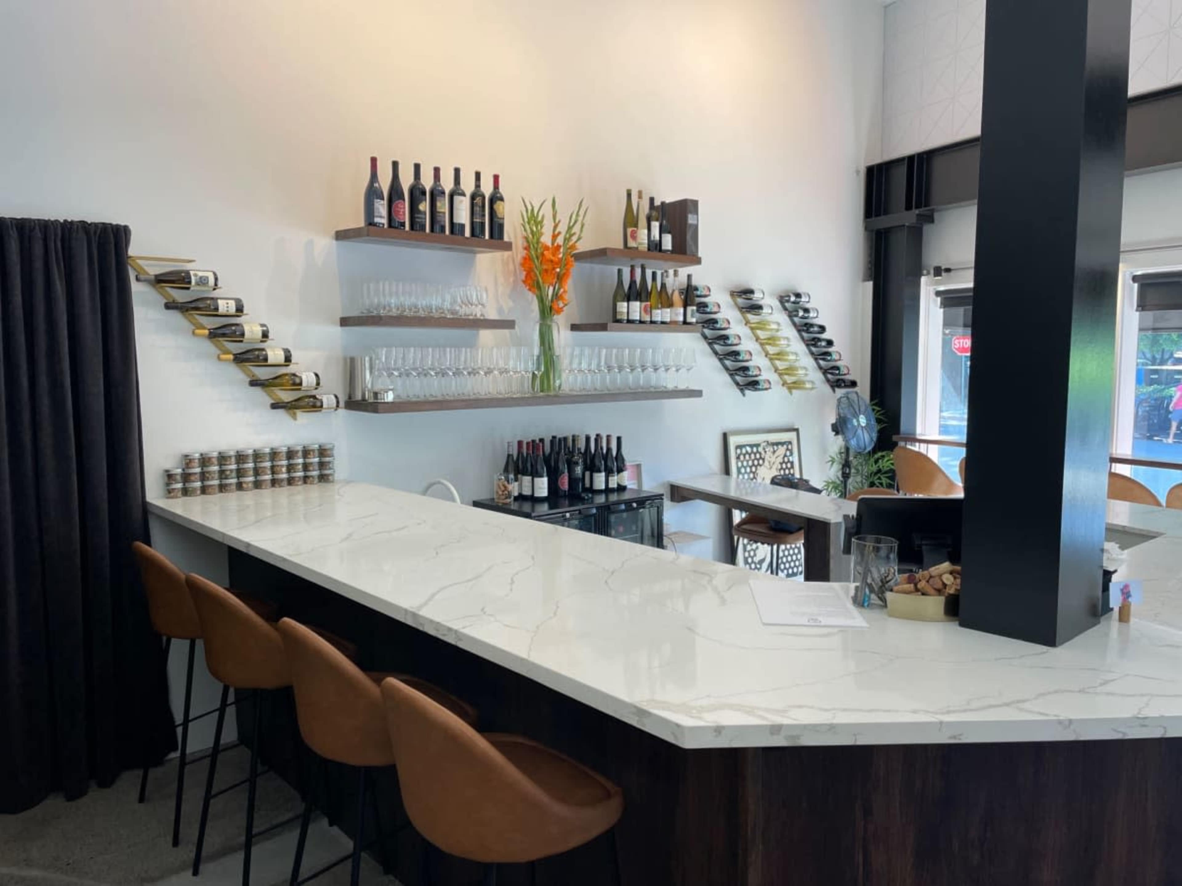The image shows a modern bar with a marble countertop, wine bottles displayed on wall-mounted racks, and a variety of glassware arranged on shelves.