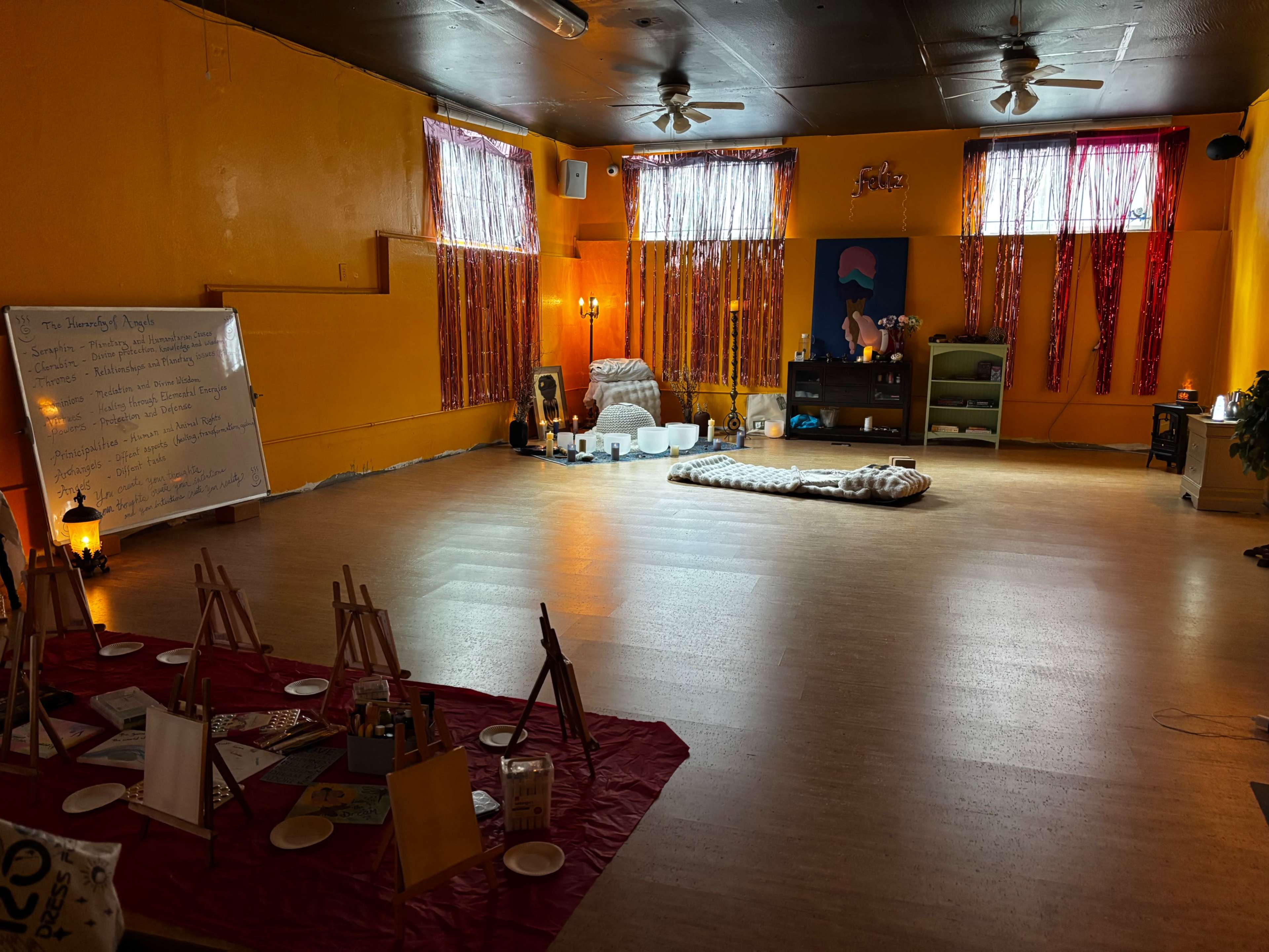 The image shows a brightly colored room with wooden flooring, featuring various items for a creative or wellness activity, including easels and a meditation area.