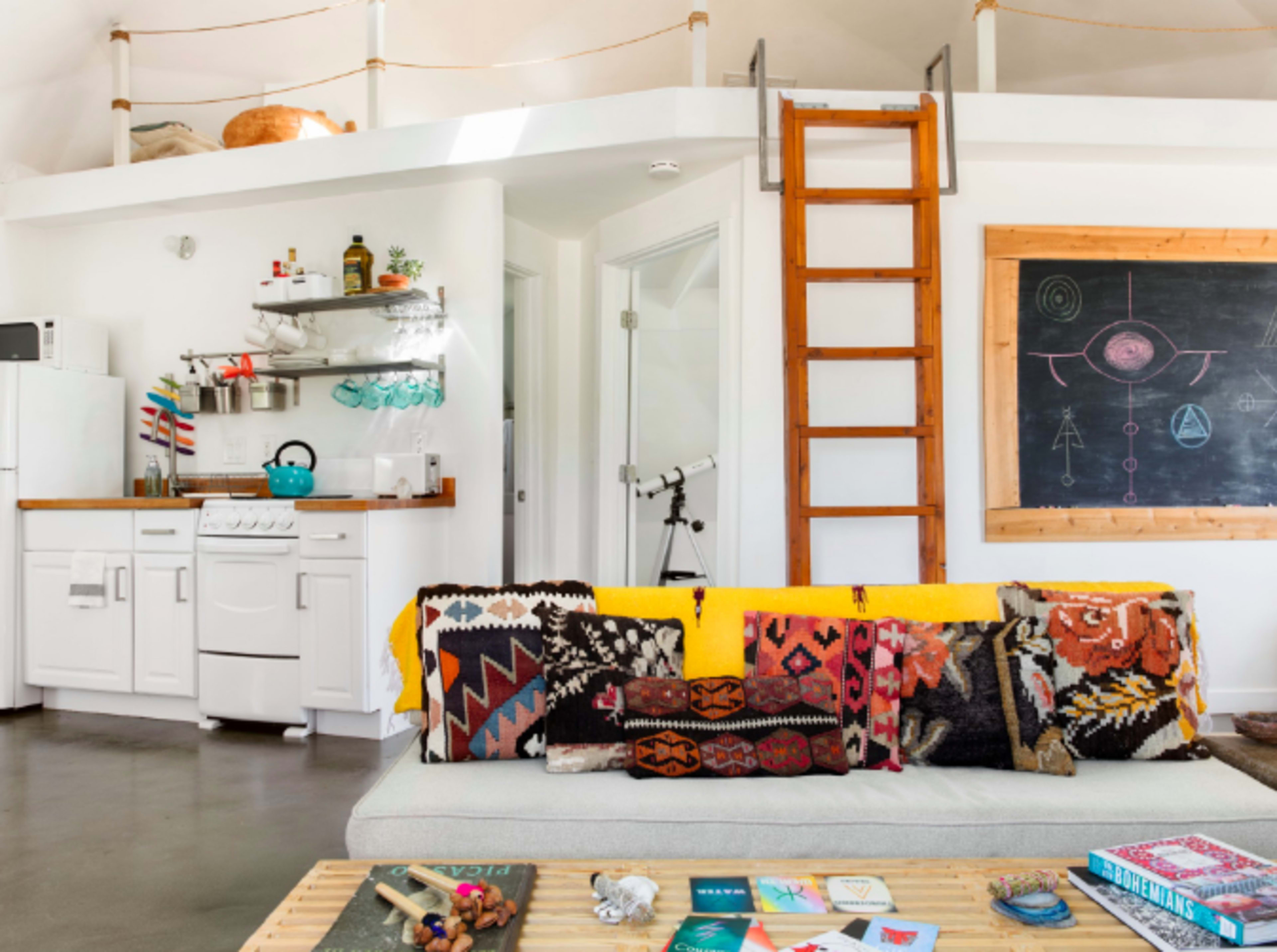 The image shows a modern living space with a kitchen area, a loft accessed by a wooden ladder, and a comfortable sofa adorned with patterned cushions.
