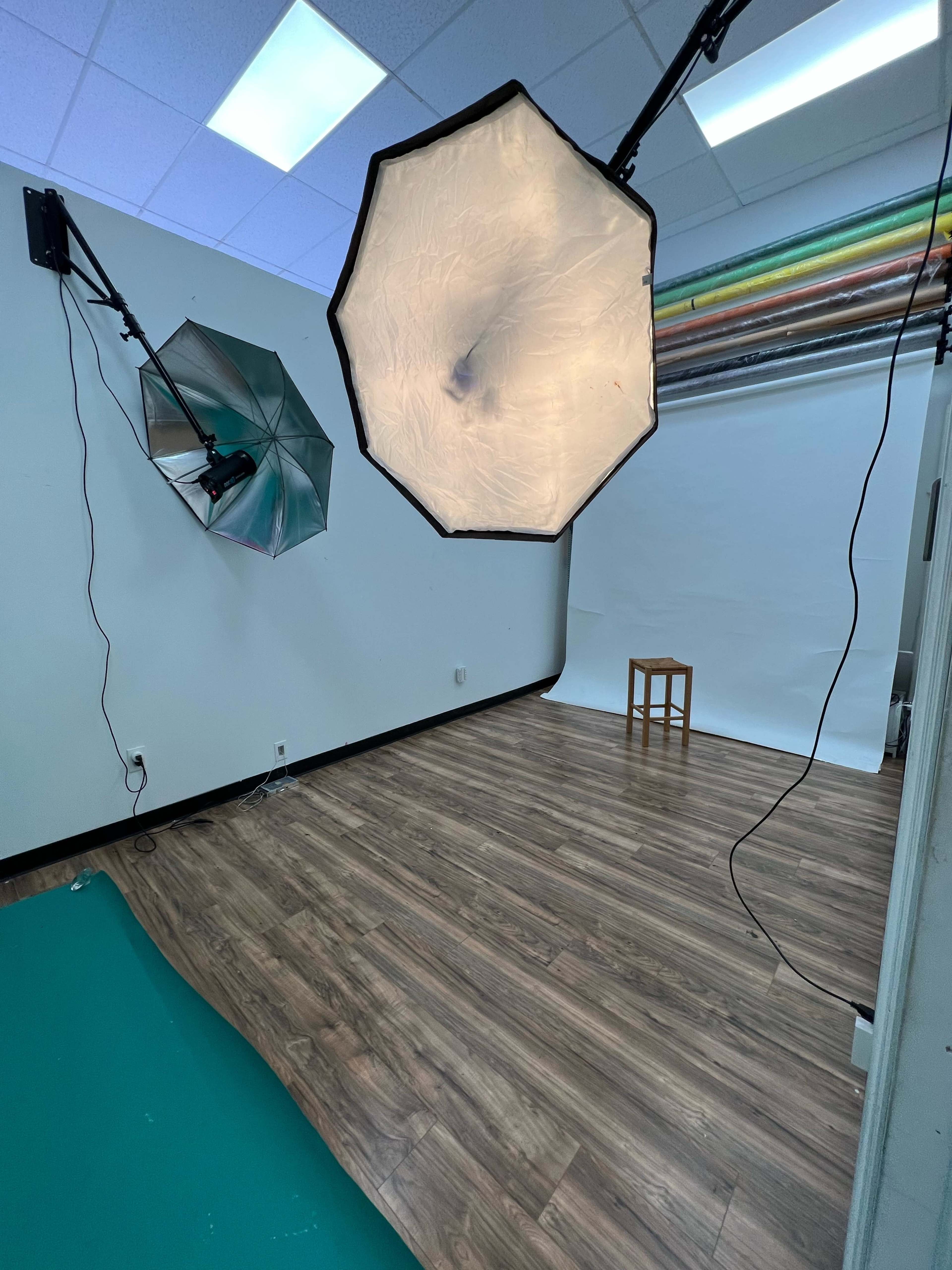 A photography studio with two light umbrellas, a white backdrop, a stool, and a green floor section.