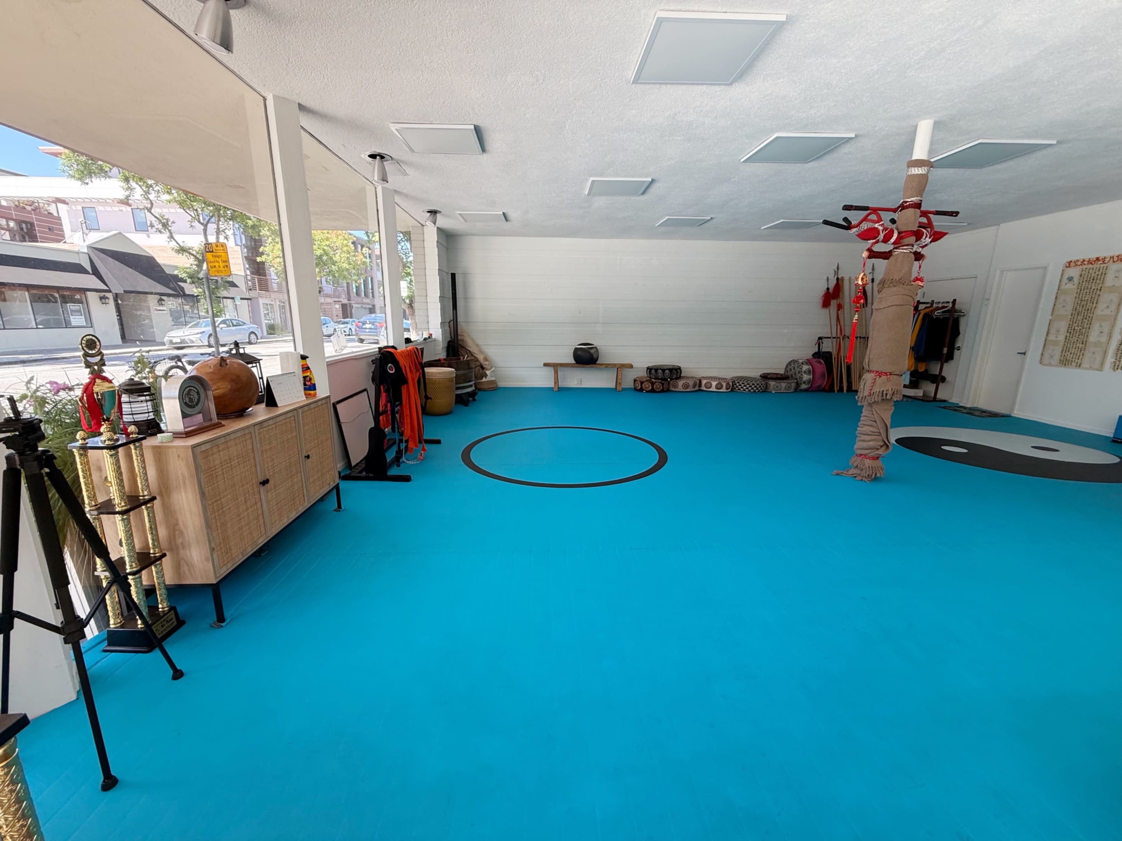 The image shows a spacious, brightly lit interior of a dojo or training hall featuring a blue rubber floor, mats, and various training equipment along the walls.