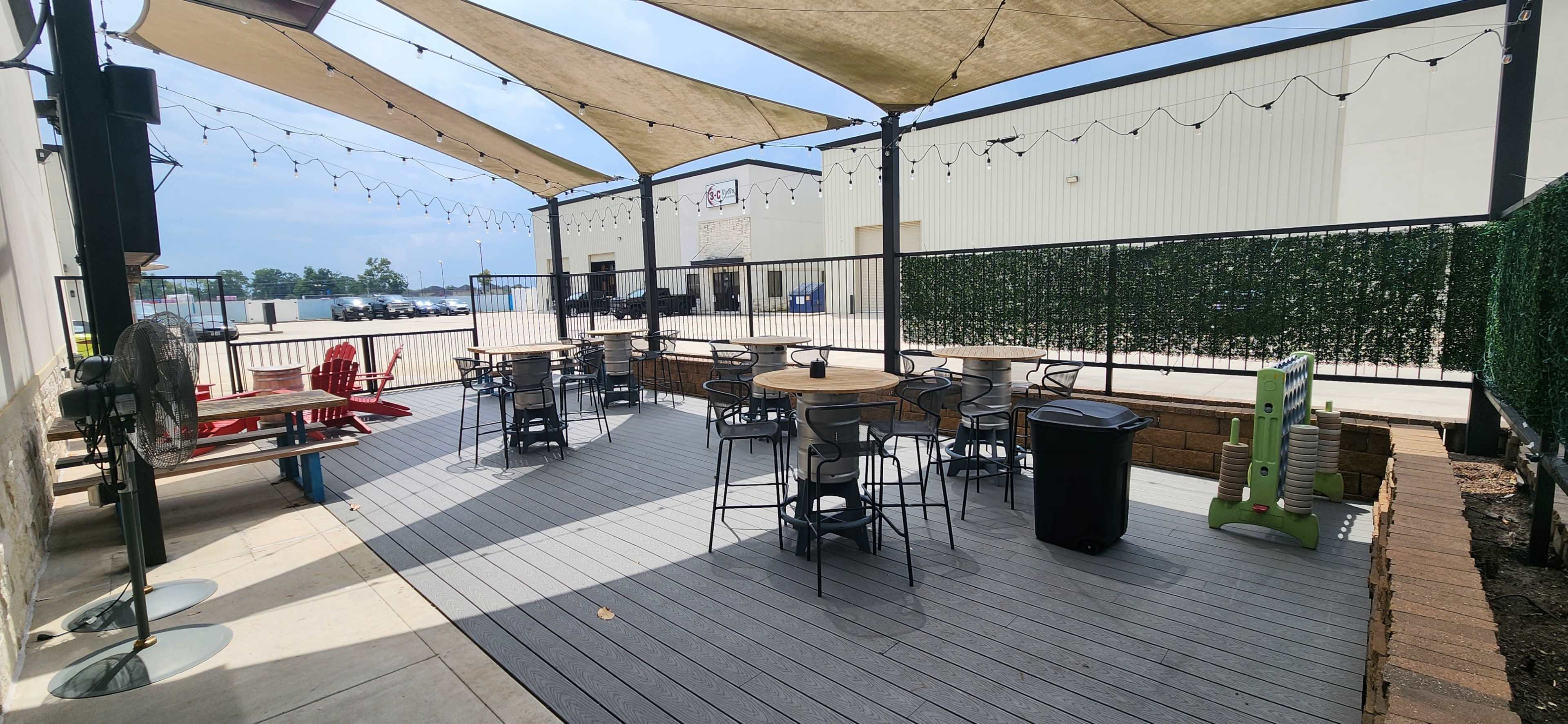 The image shows a partially covered outdoor seating area with tables and chairs on a wooden deck, surrounded by string lights and a wall of greenery.