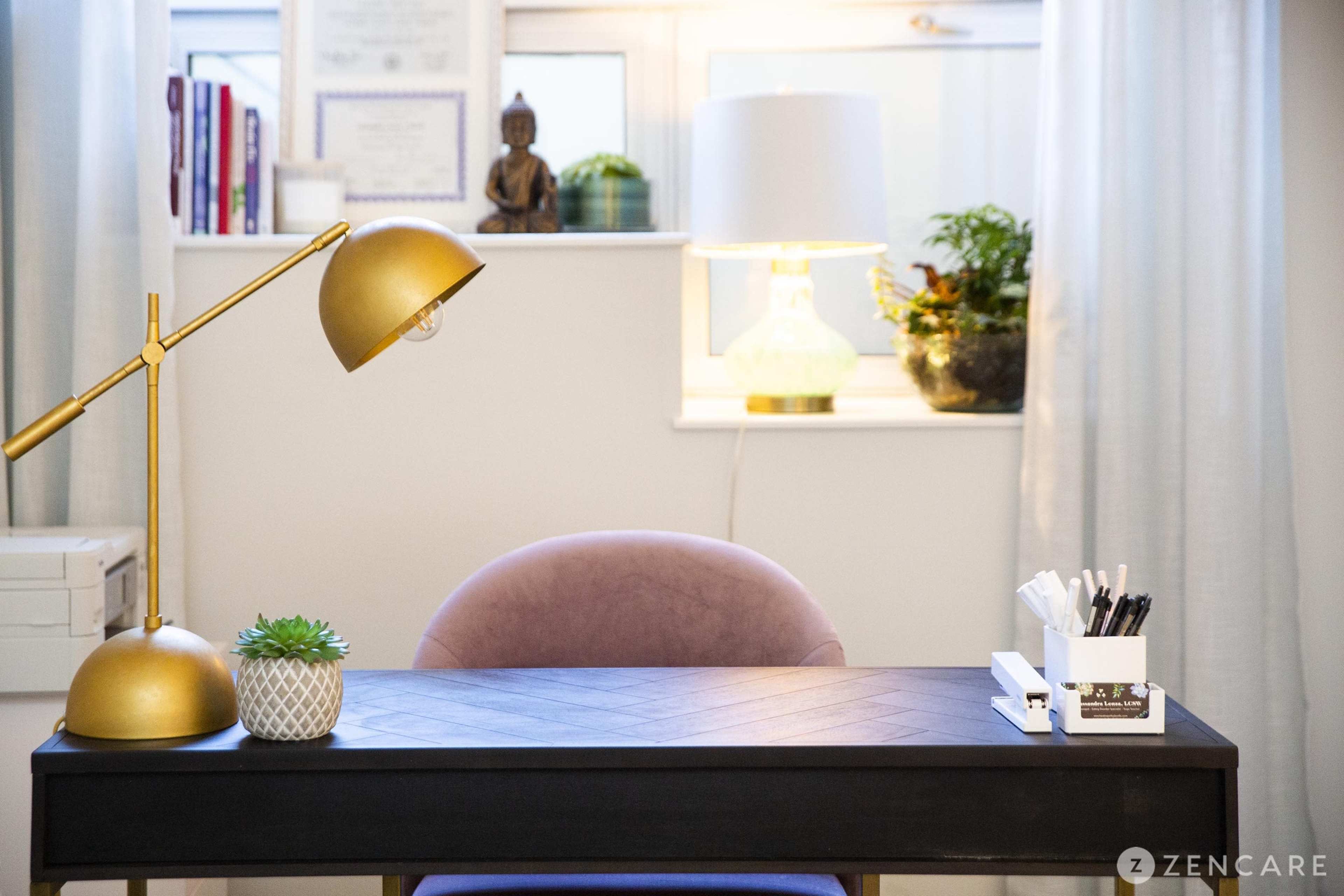 A modern desk with a pink chair, gold lamp, and organized stationery sits against a window with potted plants and framed documents in the background.