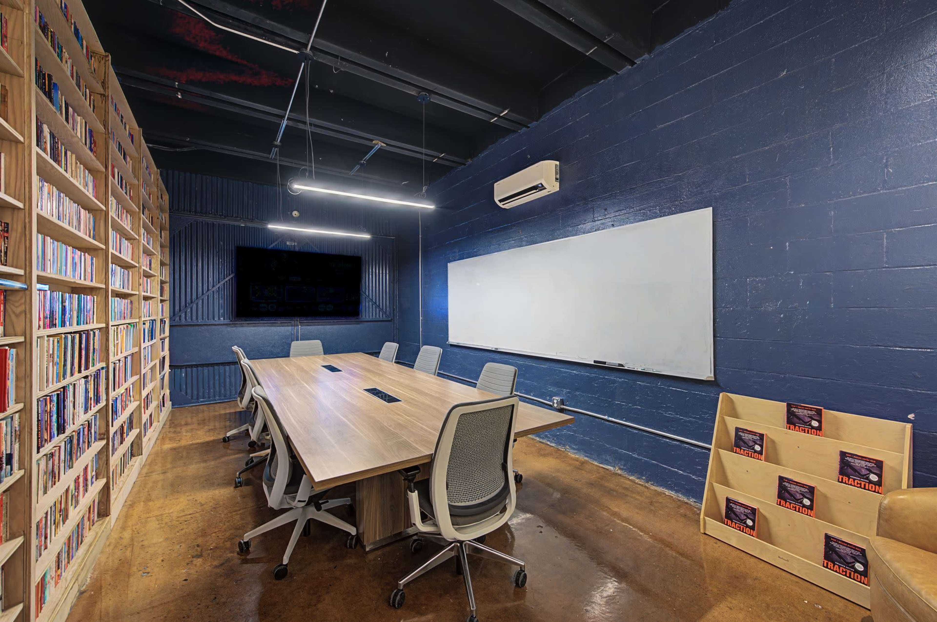 The image shows a modern conference room featuring a large table surrounded by chairs, a whiteboard mounted on the wall, and a bookshelf filled with books.