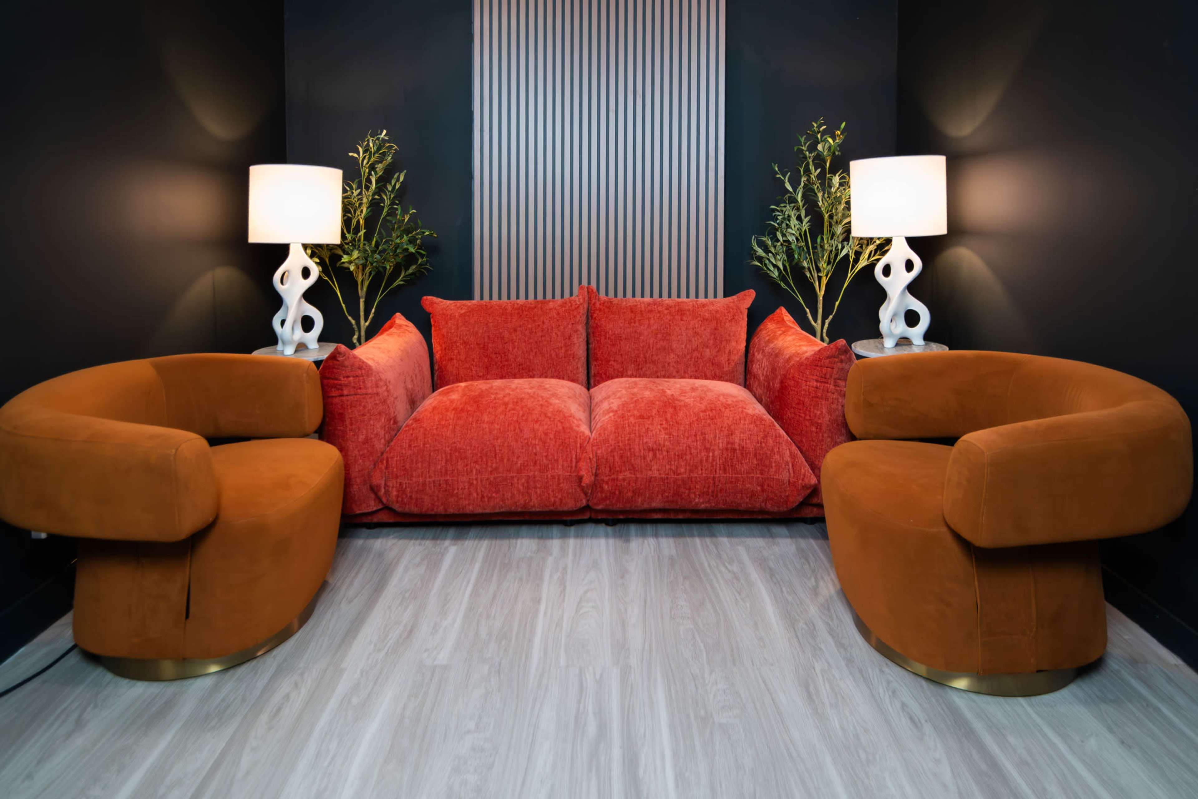 The image shows a modern living room setup featuring a red plush sofa flanked by two curved brown chairs, with two table lamps and potted plants against a dark wall.
