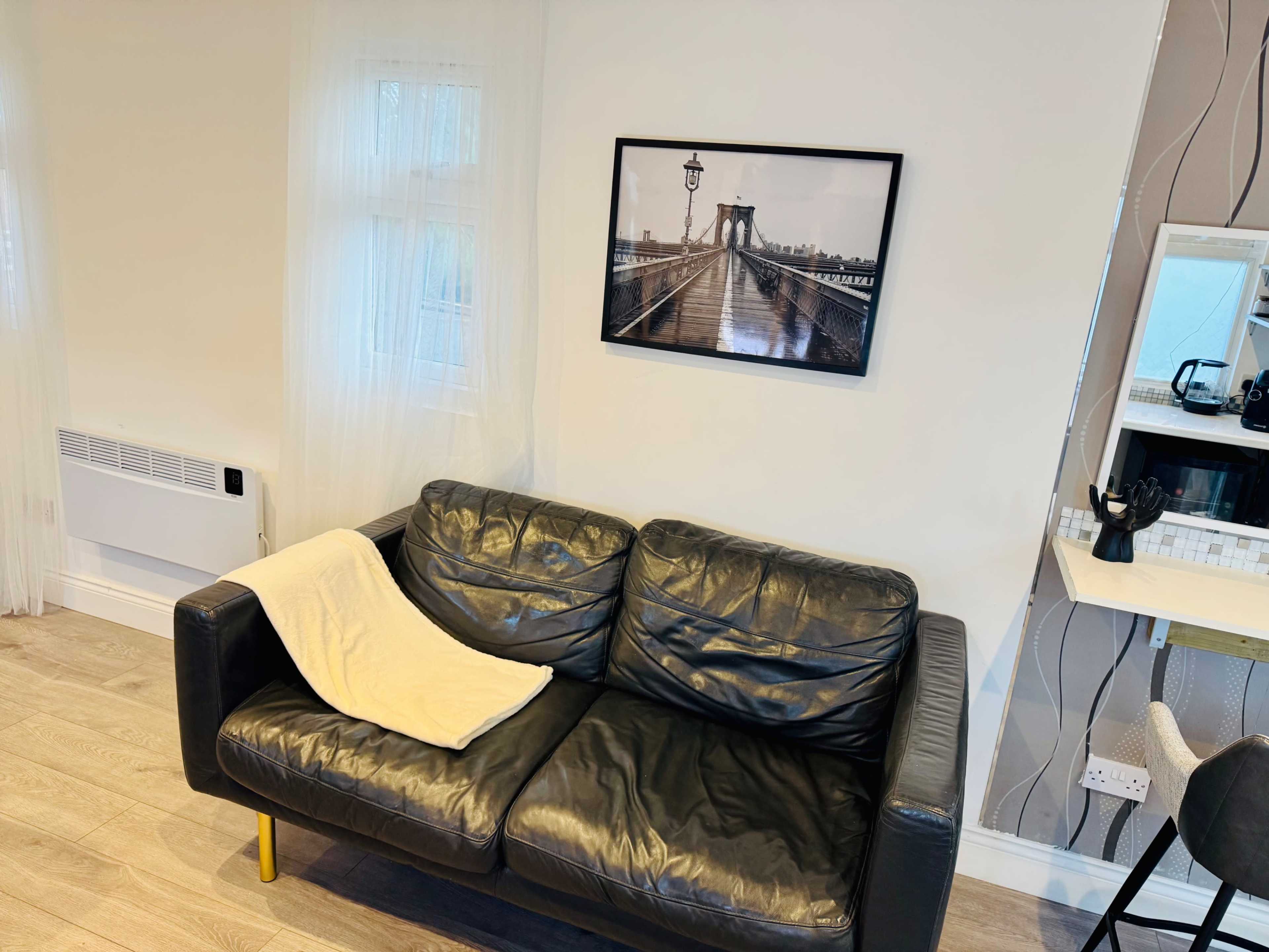 A black leather sofa with a white throw draped over it is positioned against a light-colored wall, beside a framed black-and-white photograph of a bridge.