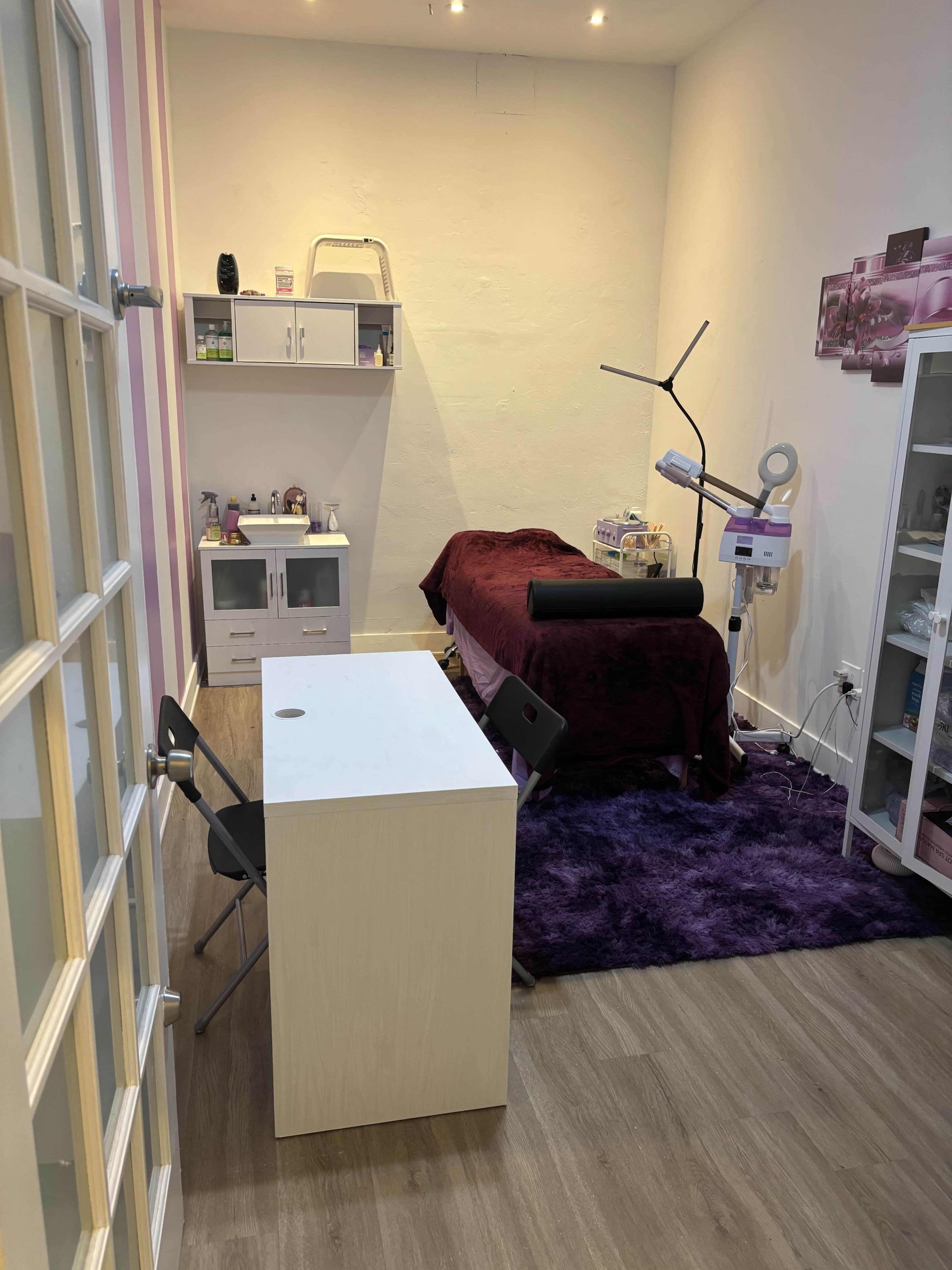 The image shows a small treatment room with a bed covered in a dark blanket, a desk with two black chairs, and various beauty equipment on shelves.