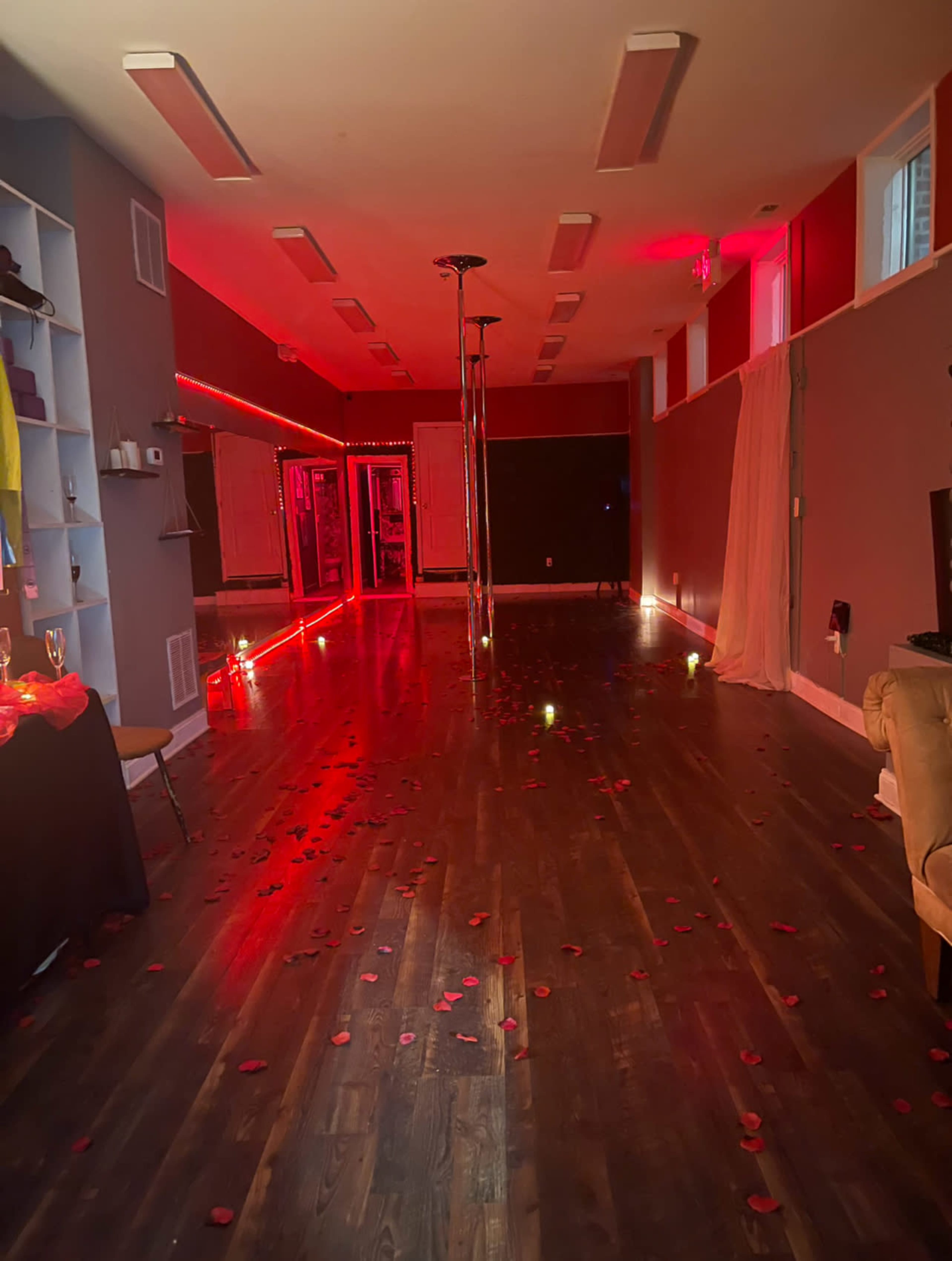 The interior of a dimly lit room features a wooden floor scattered with rose petals, a pole in the center, and red lighting along the walls.