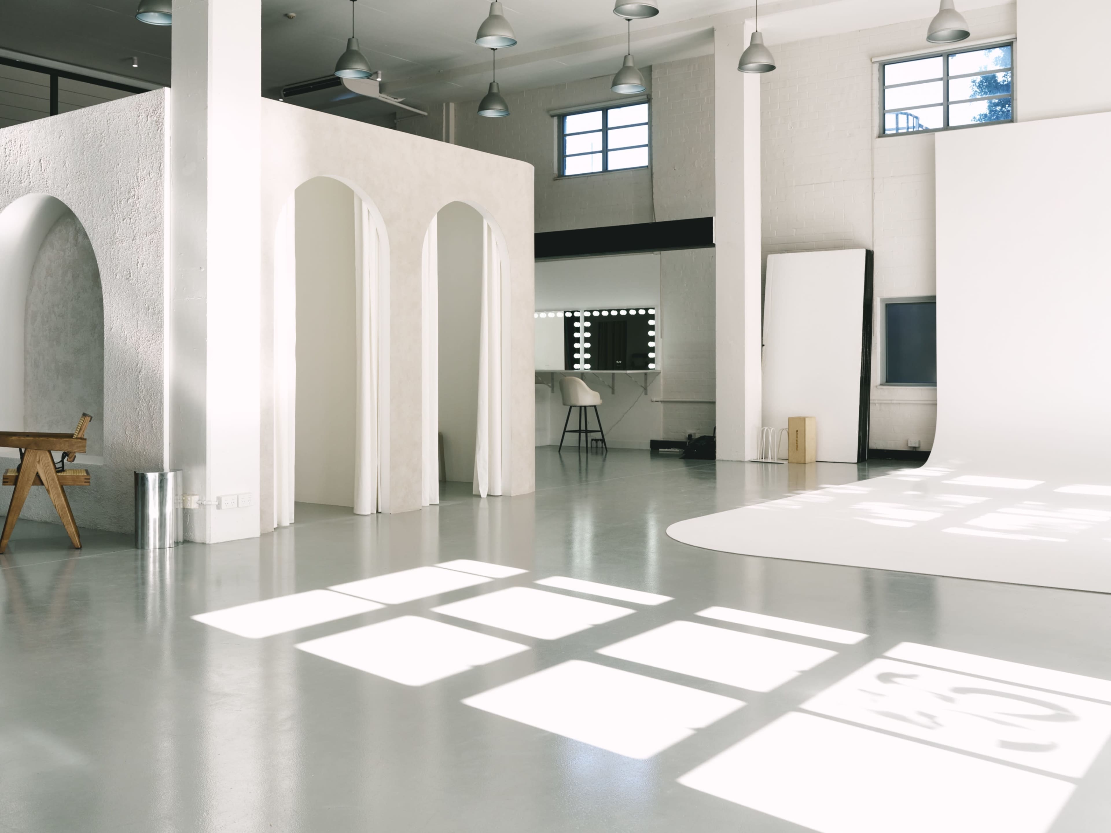 The image shows a spacious, well-lit studio with white walls, large windows, and various architectural elements, including arches and a white backdrop on the floor.