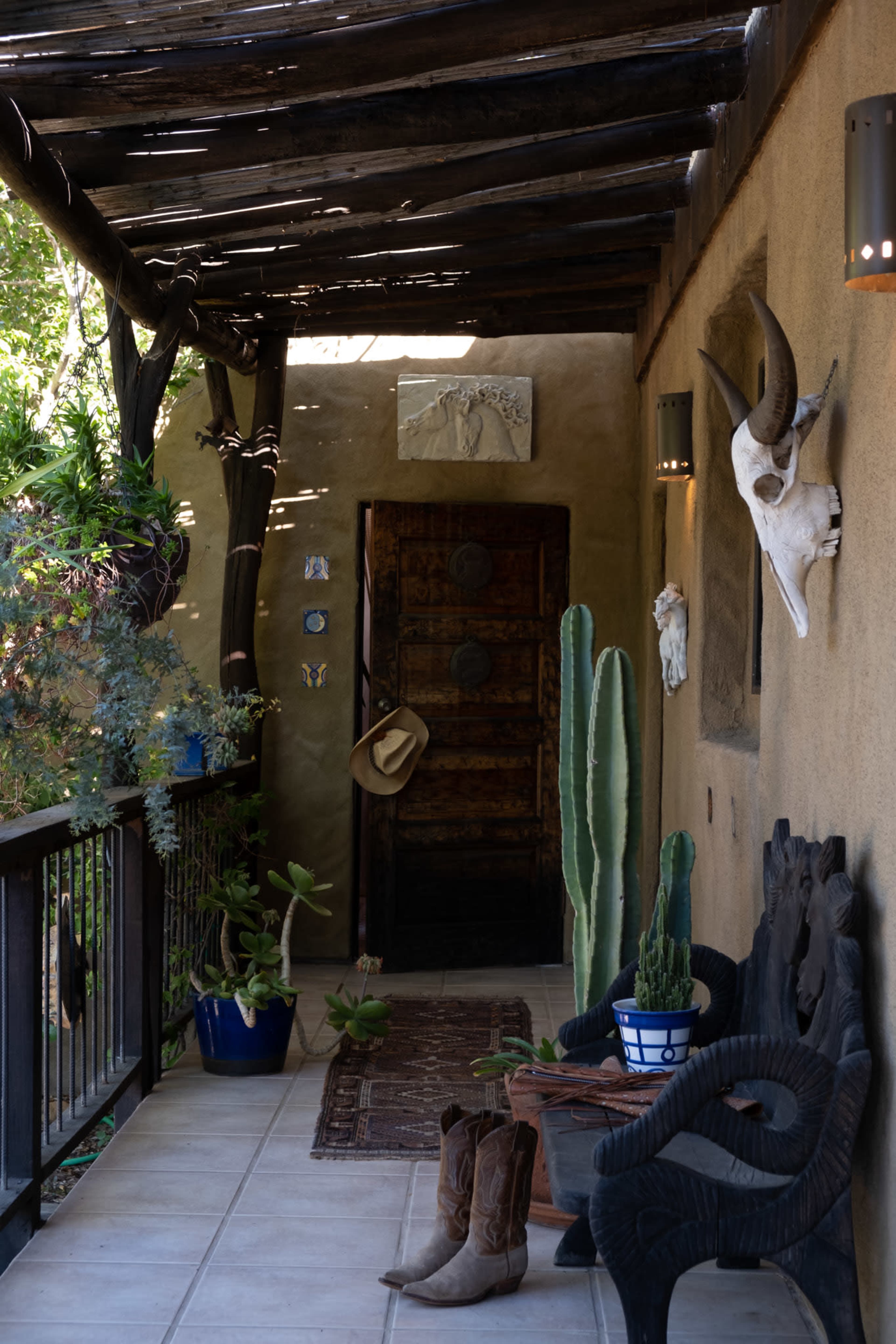 A rustic porch with a wooden ceiling, a large door, cacti, decorative animal skulls, and a pair of cowboy boots near a carved wooden chair.