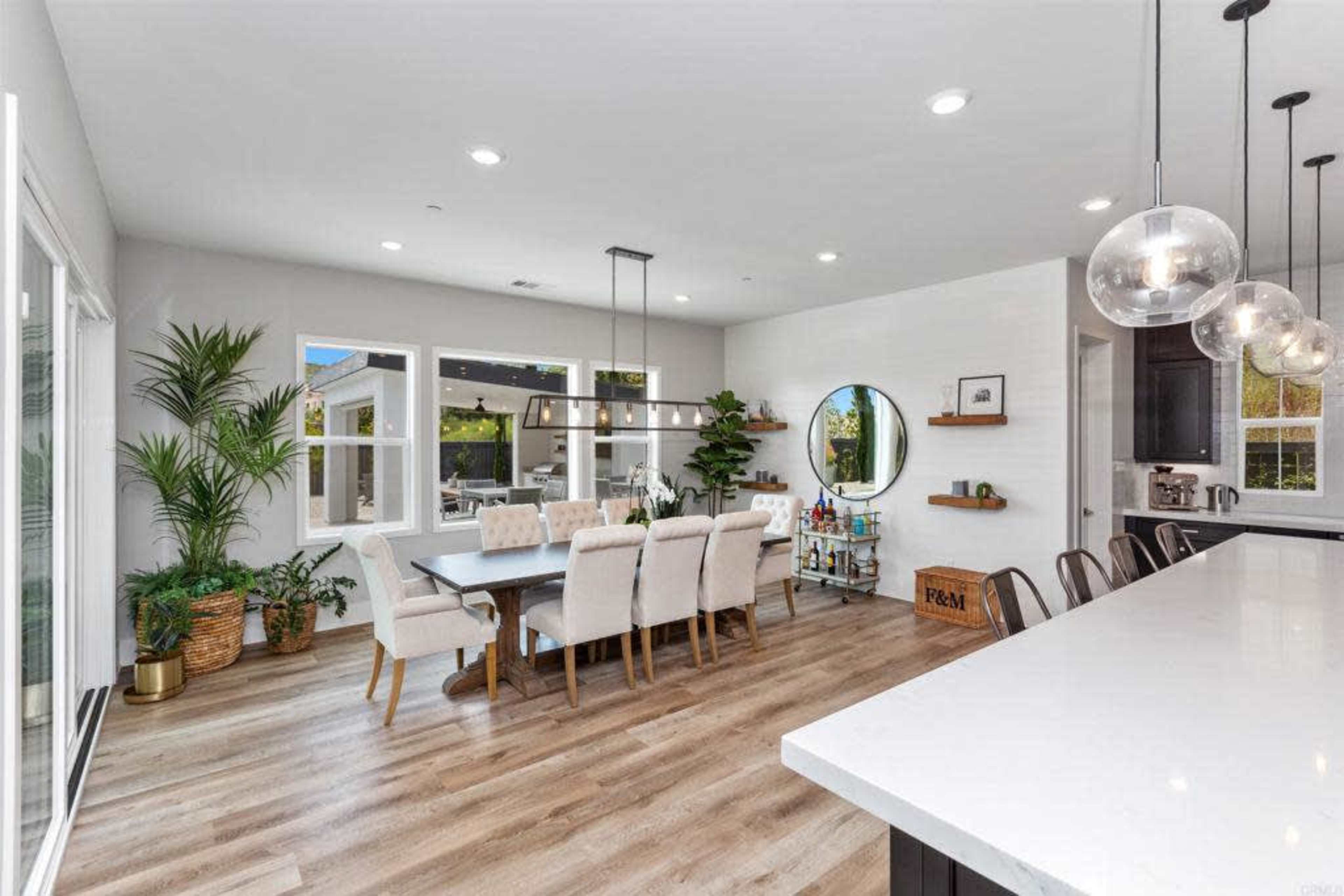 A modern dining area with a large table surrounded by upholstered chairs, large windows, and a circular mirror on the wall.