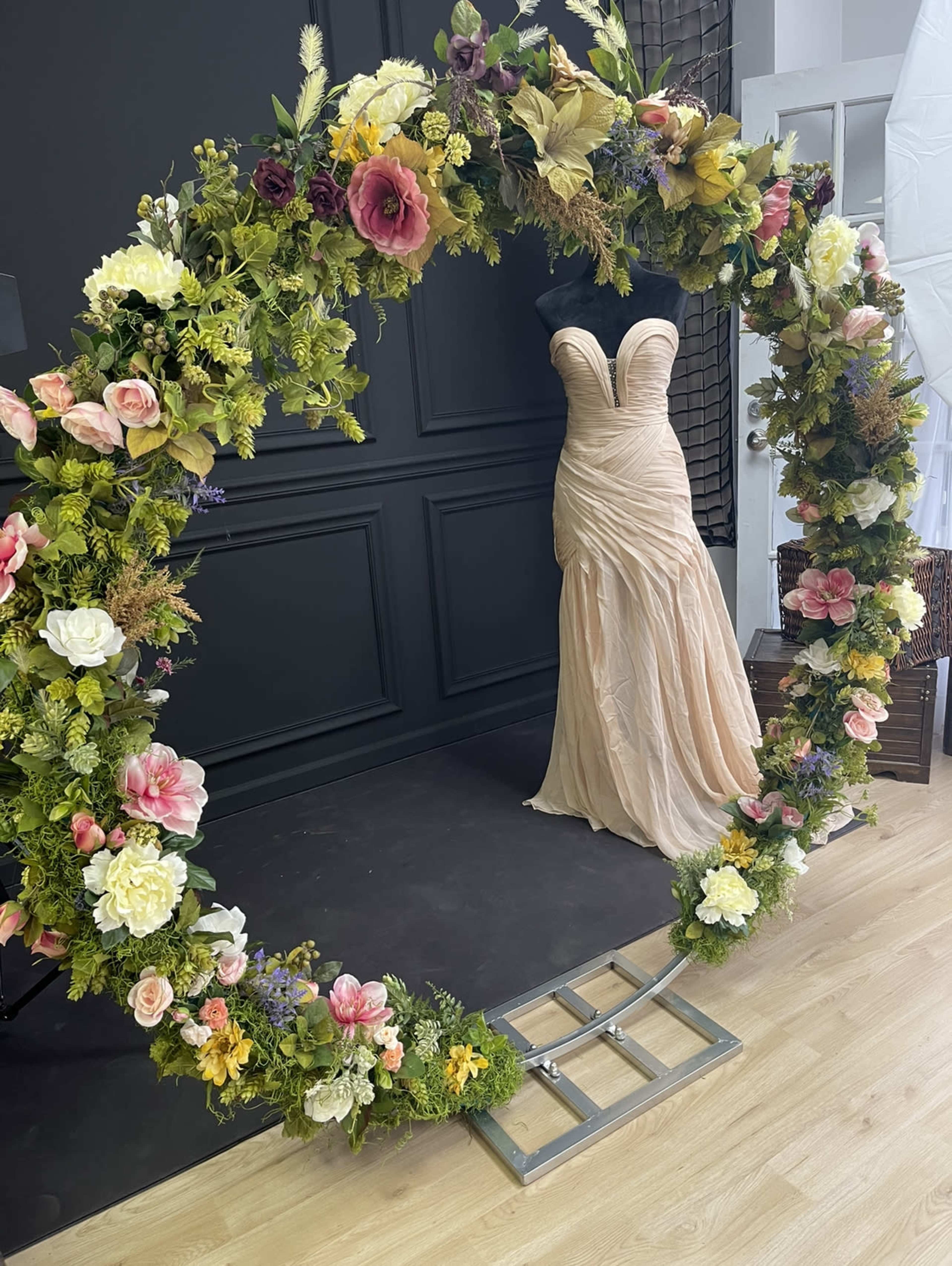 A floral arch surrounds a formal gown displayed on a mannequin against a dark wall.
