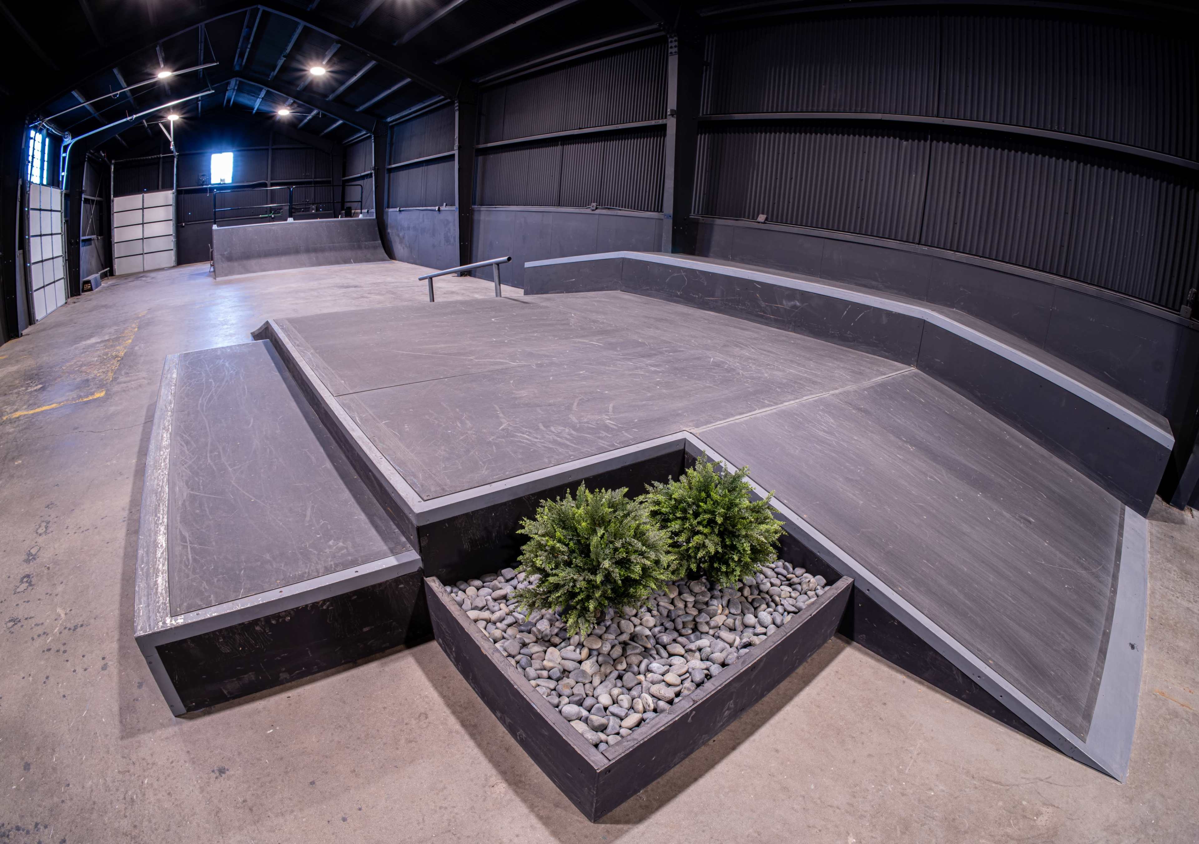 The image shows a spacious indoor skatepark featuring various ramps, rails, and a planter area with rocks and greenery.