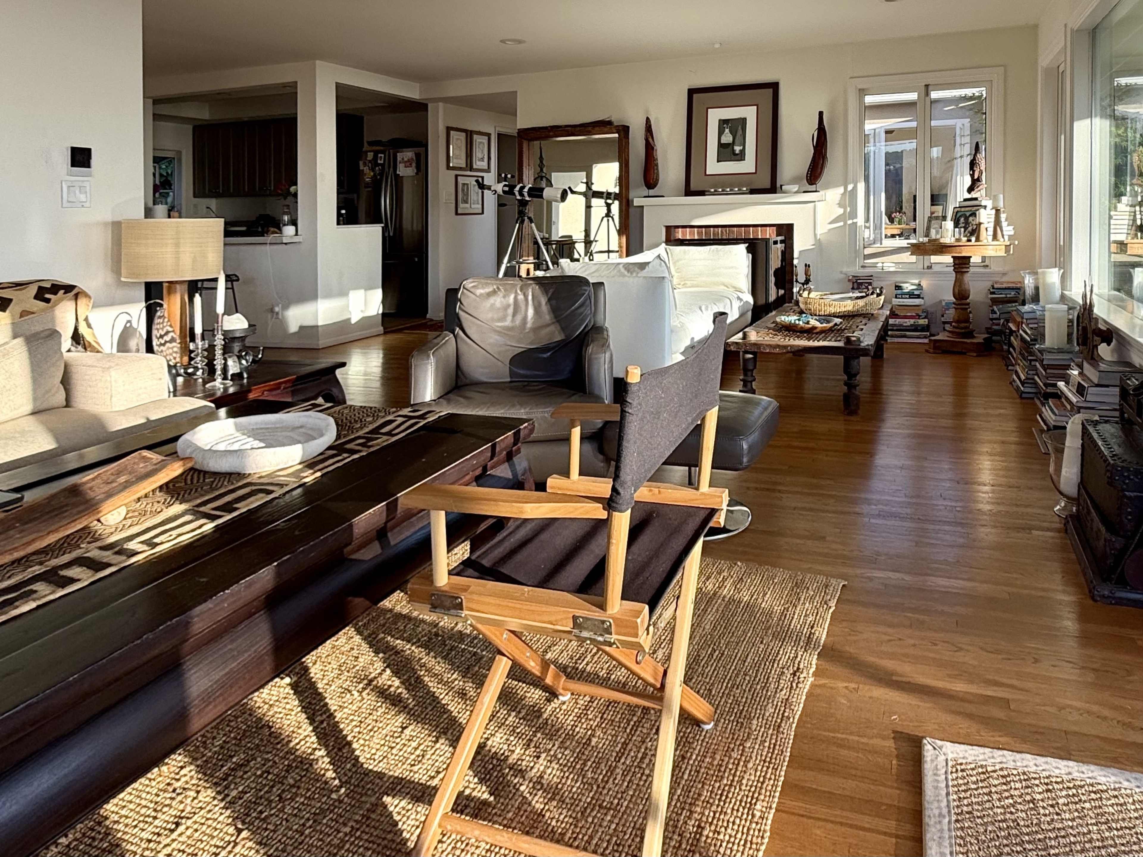 A spacious living room features a mix of furniture, including a director's chair, a coffee table, and various decorations, with sunlight streaming through large windows.