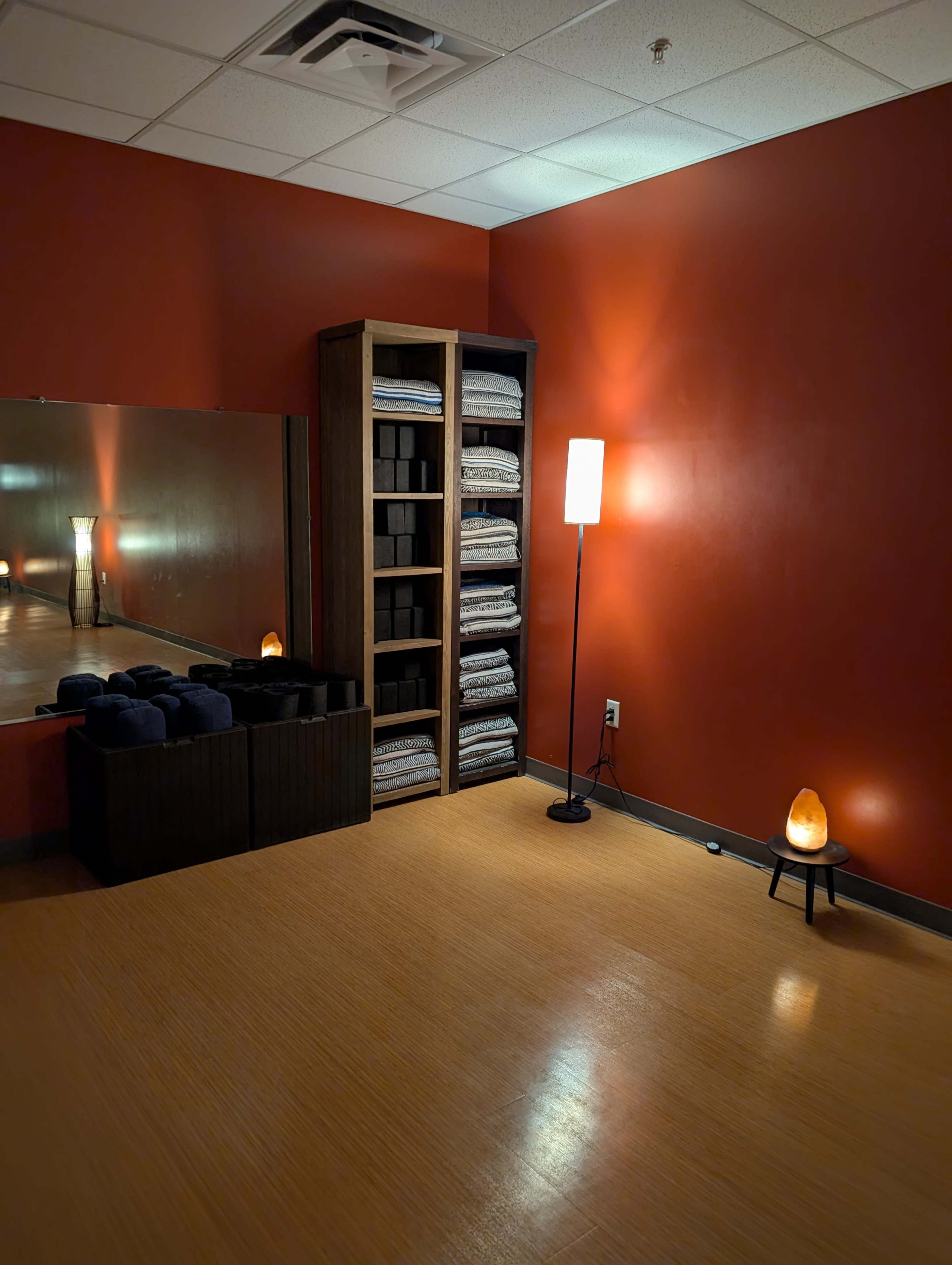A calming interior features a shelf with neatly stacked towels, a lamp, a salt lamp, and a large mirror against a warm red wall.