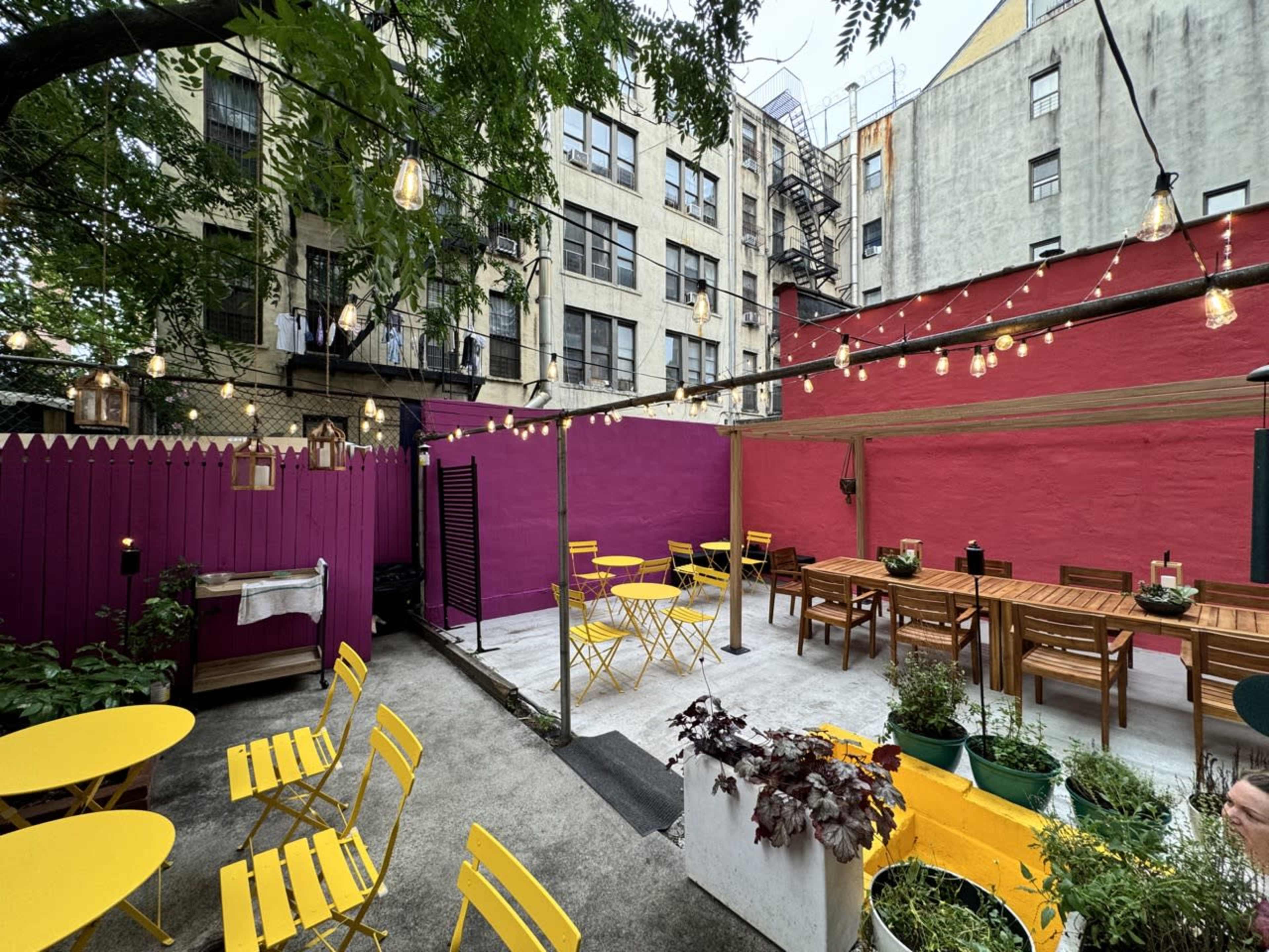 A colorful outdoor dining area with yellow chairs and wooden tables, surrounded by a purple and red wall and decorated with string lights.