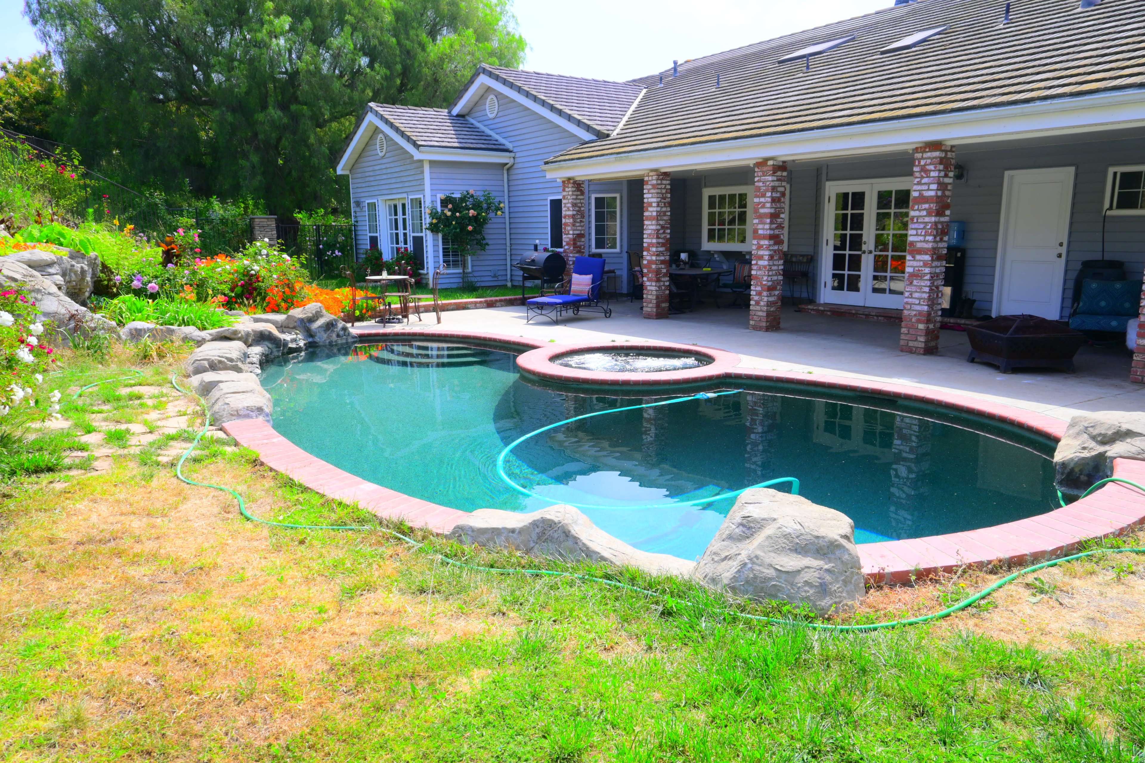 A backyard with a landscaped area featuring a swimming pool surrounded by rocks and grass, adjacent to a house with a porch.