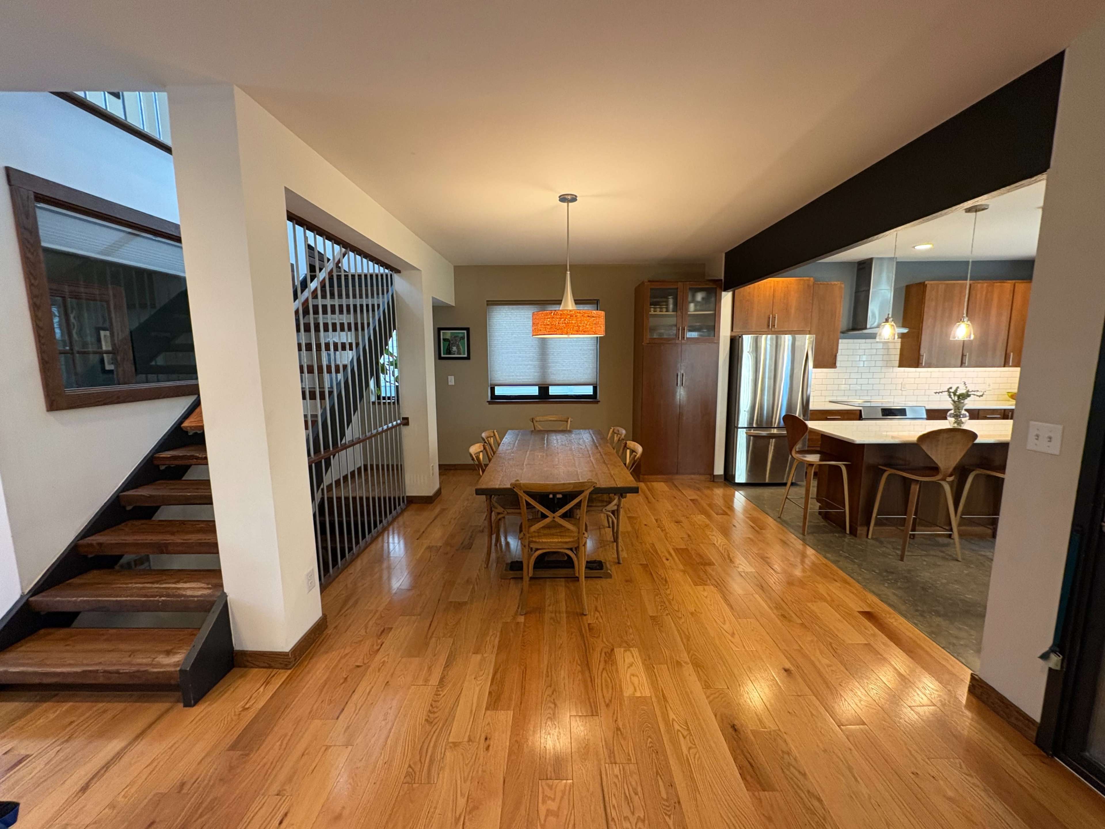 The image shows a modern dining area with a wooden table, surrounded by chairs, and an open kitchen in the background, featuring stainless steel appliances and a spiral staircase nearby.