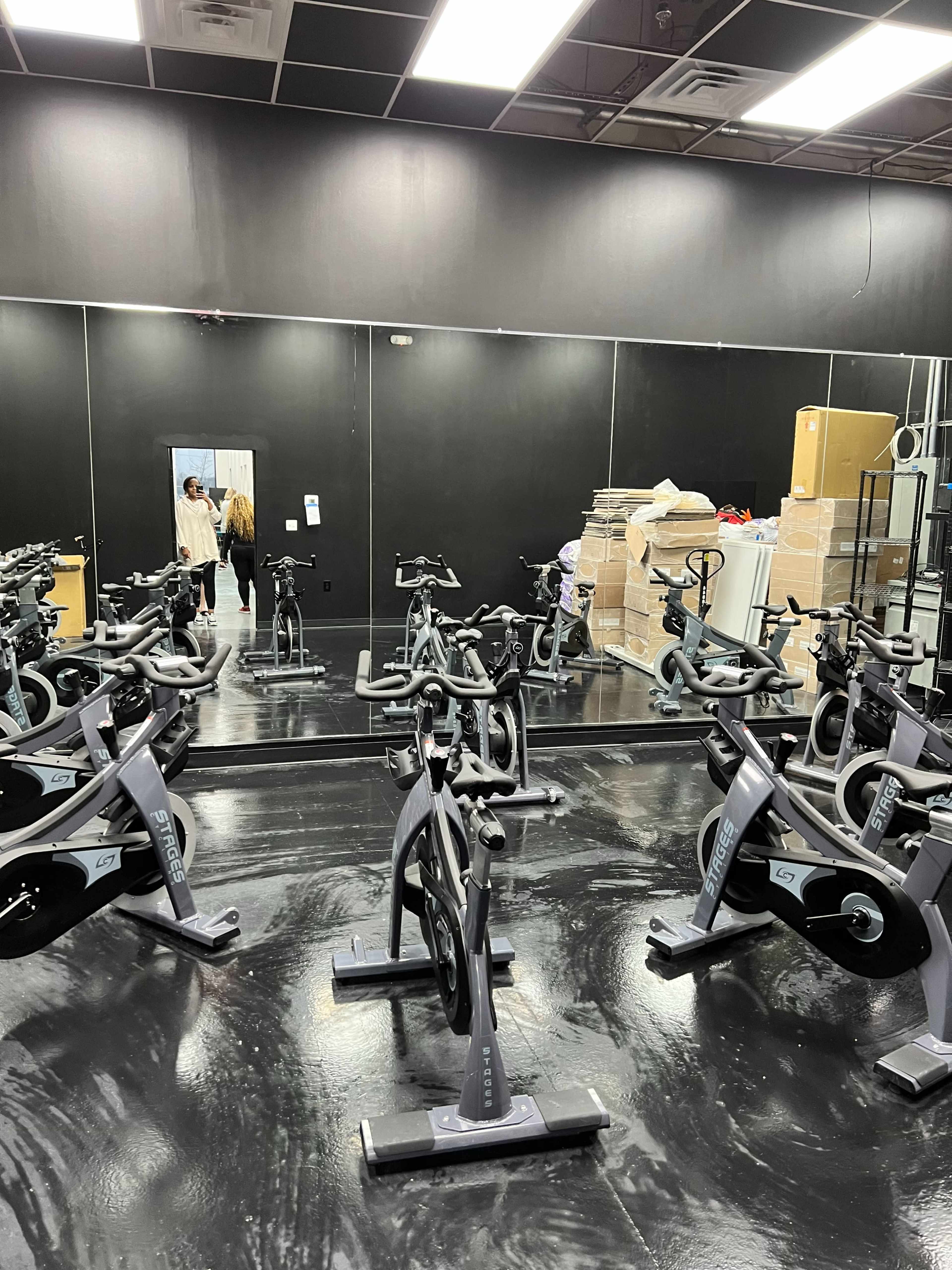 A spacious indoor cycling room with several stationary bikes and a large mirror reflecting the entrance and boxes stacked in the corner.