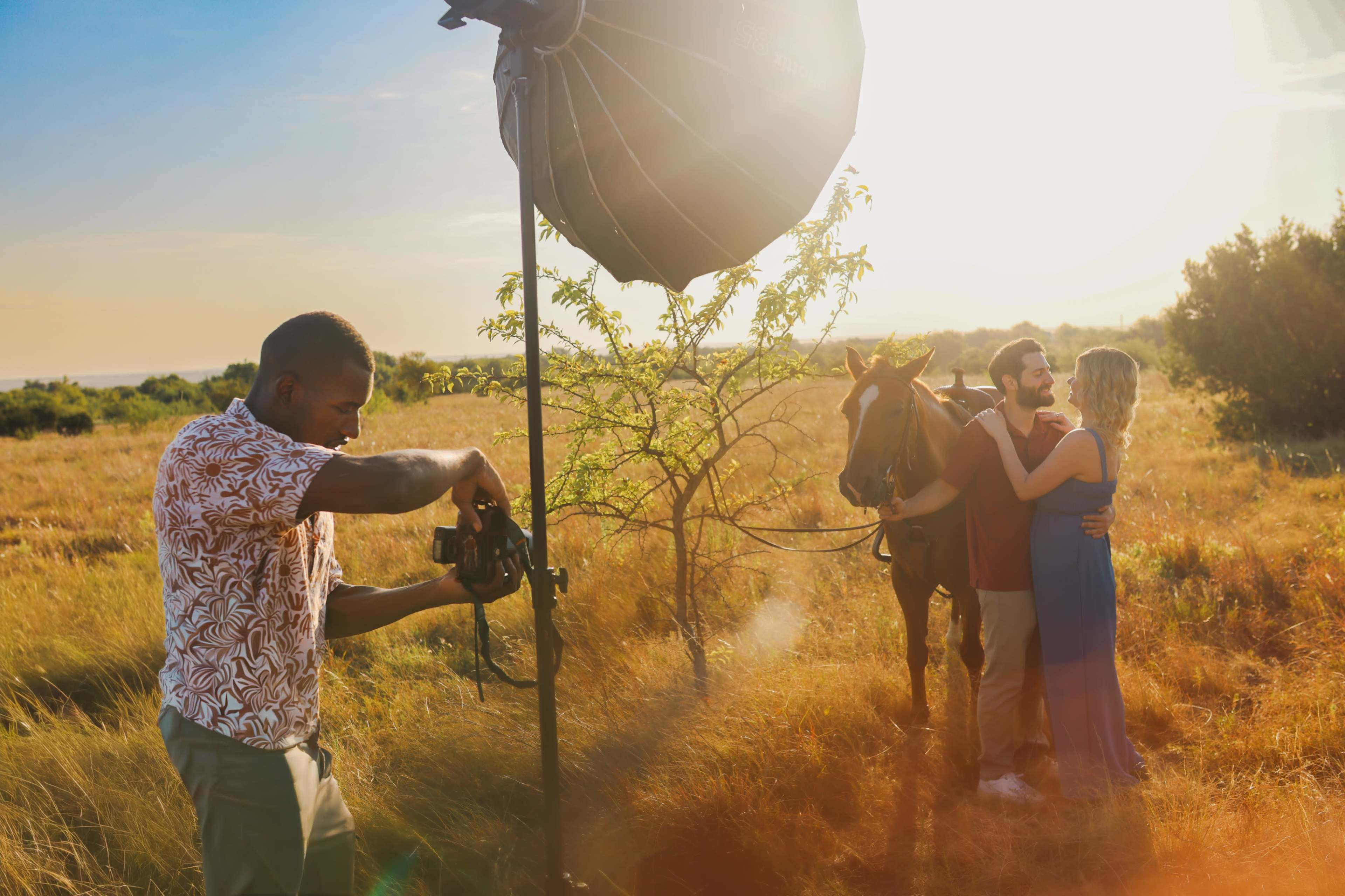A photographer captures a couple standing next to a horse in a golden field during sunset.