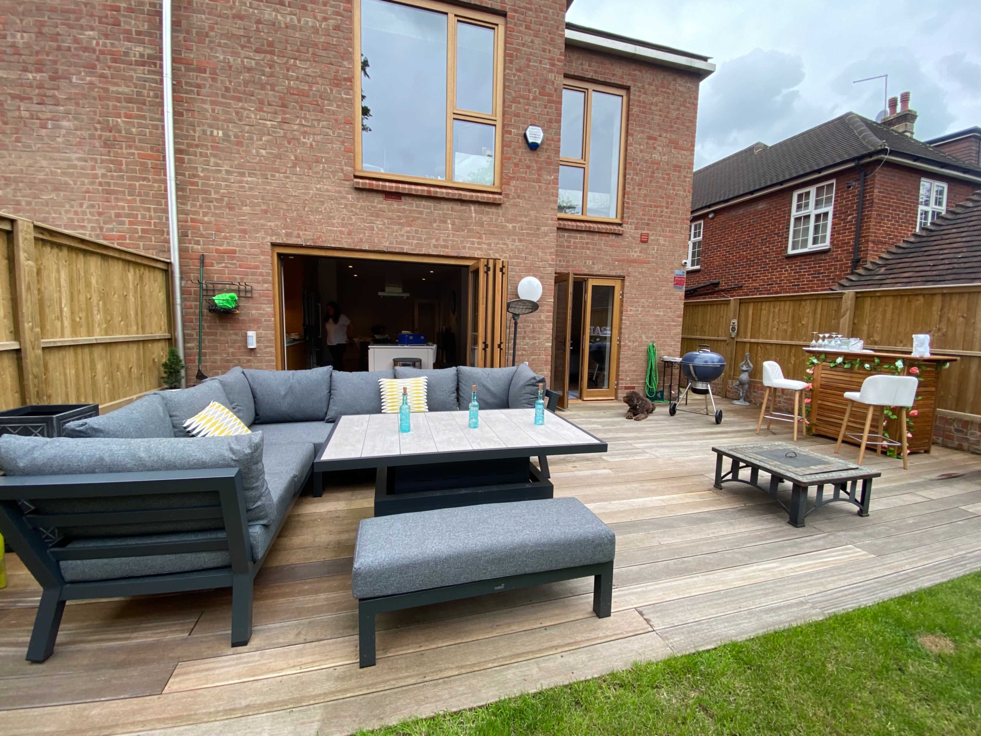 A backyard patio with a large gray sectional sofa, a table with drinks, and a grill, adjacent to a brick house.