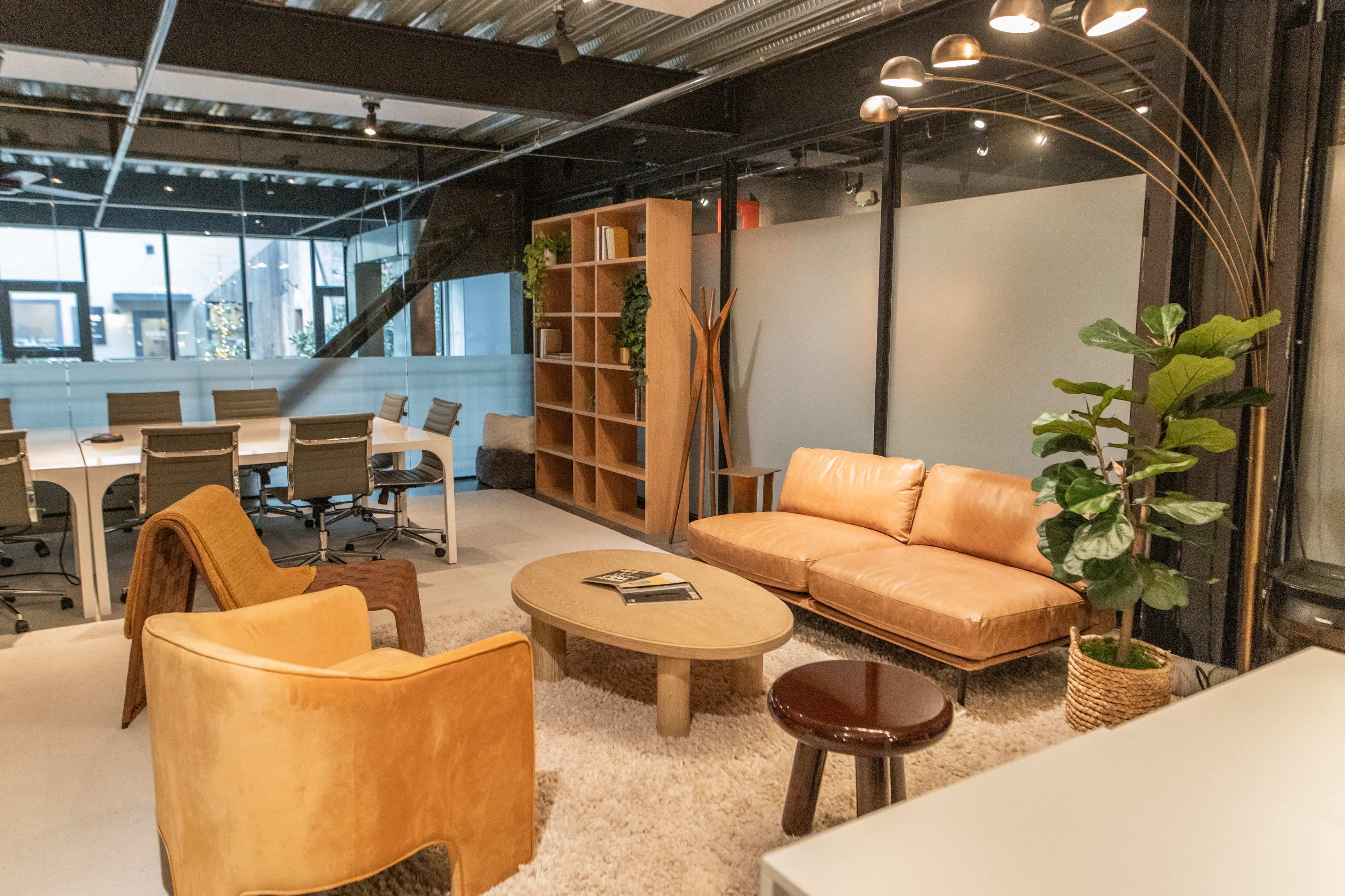 The image shows a modern office space featuring a cozy seating area with a tan sofa, a round coffee table, and a green potted plant, alongside a meeting room visible in the background.